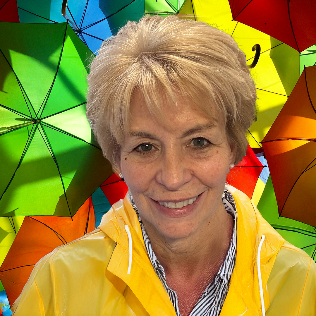 A smiling woman with short blonde hair wearing a yellow raincoat, surrounded by colorful open umbrellas in green, blue, yellow, orange, and red.
