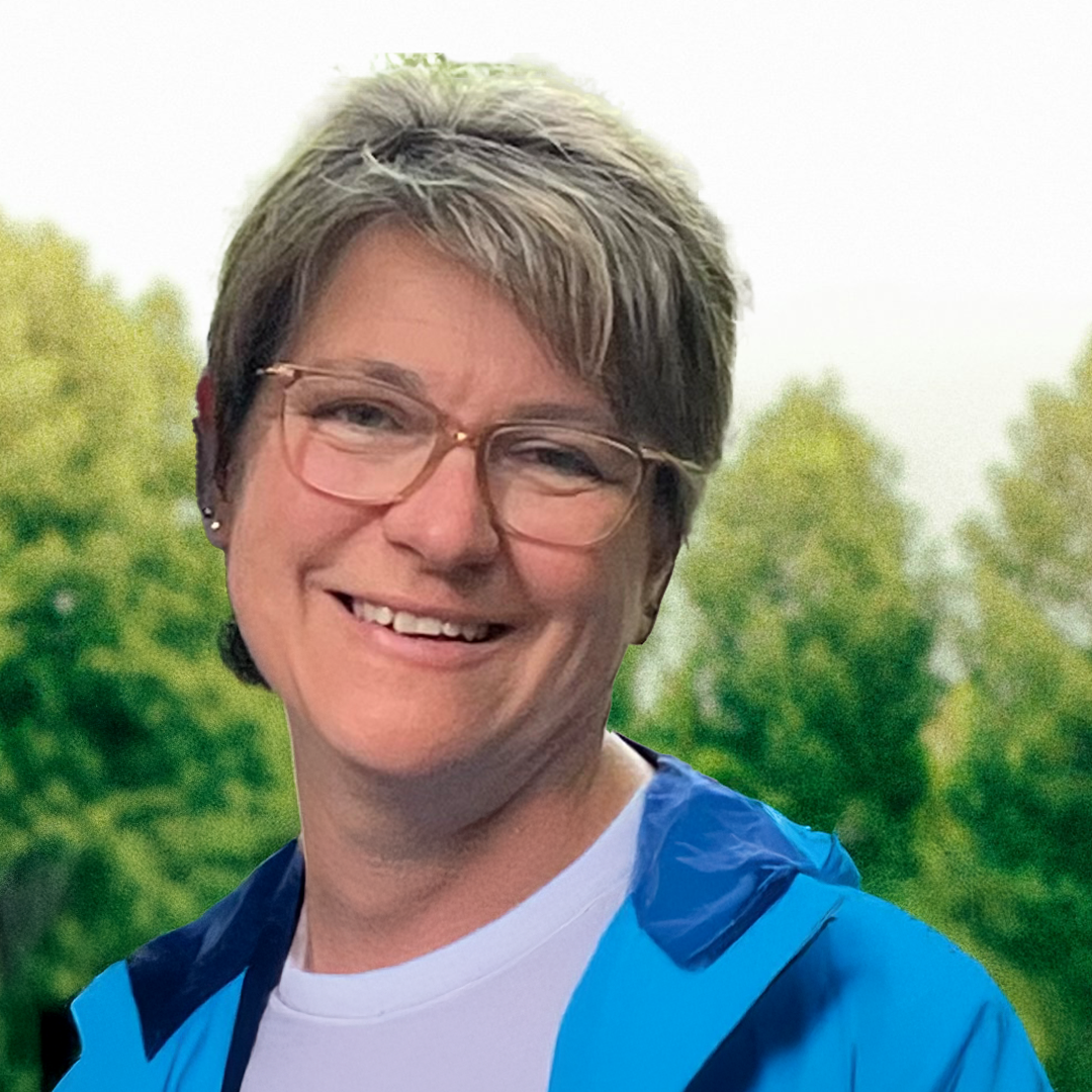 Smiling woman with short light brown hair, glasses, and a blue jacket outdoors with green trees in the background.