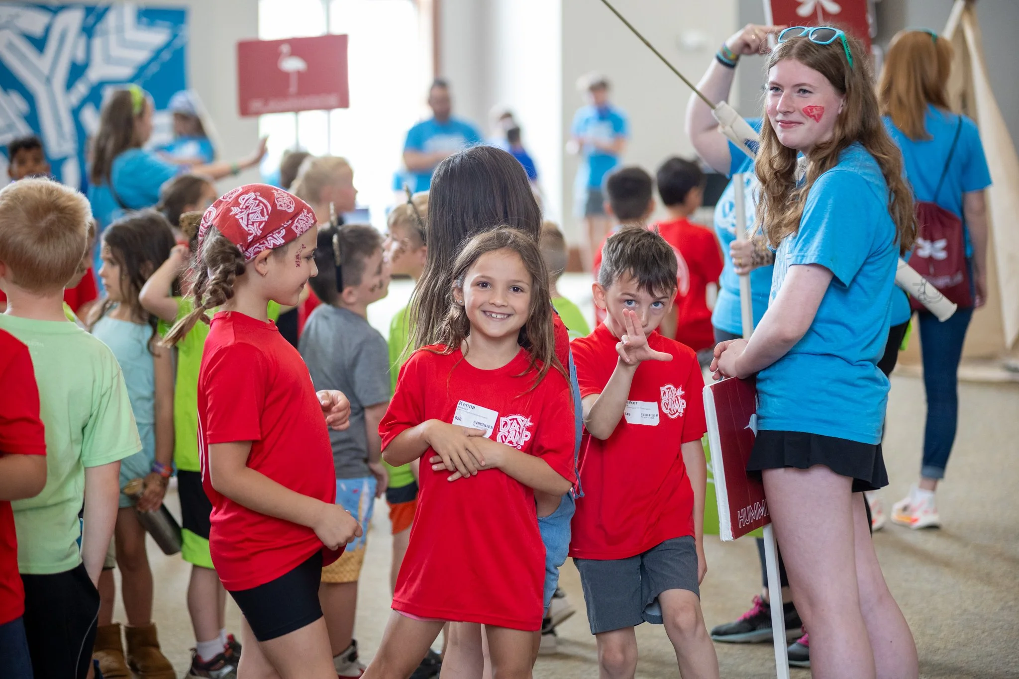 Children at a summer camp, some in red shirts, and camp counselors in blue shirts, engaging and smiling indoors.