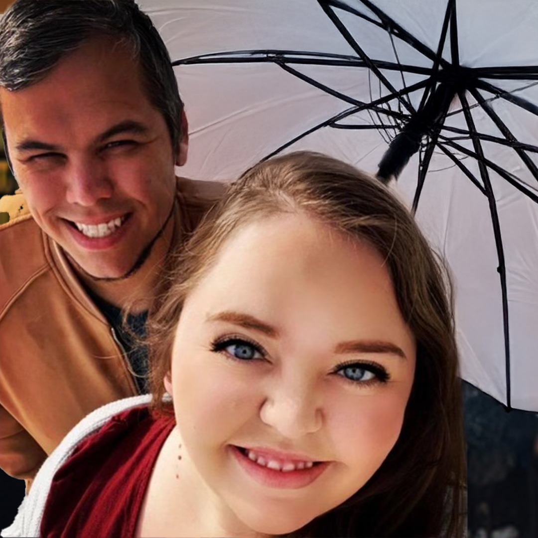 A man and a woman taking a selfie outdoors under a black and white umbrella. The man is smiling broadly, showing his teeth, and has short dark hair. The woman has blue eyes, long brown hair, and a big smile. It appears to be rainy or wet outside.