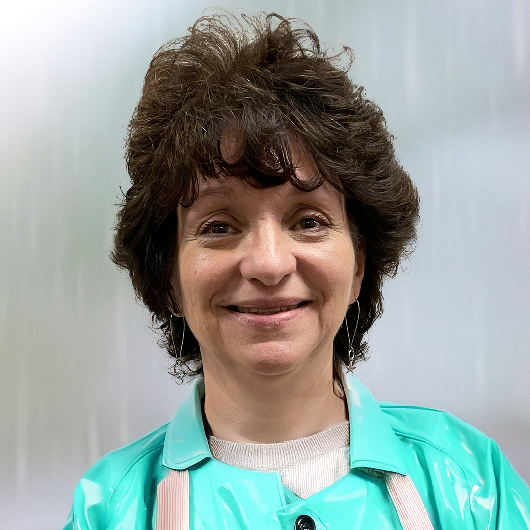 A woman smiling, with short curly brown hair, wearing a light blue coat and earrings, standing against a plain background.