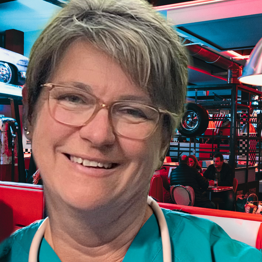 Smiling woman with short light brown hair, glasses, and a blue jacket outdoors with green trees in the background.
