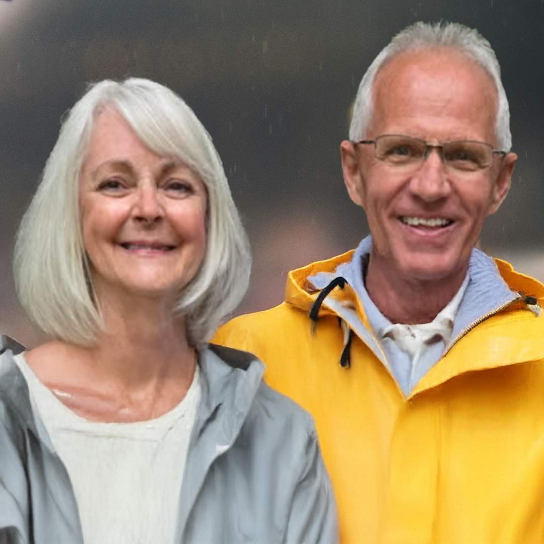 Smiling elderly couple outdoors, woman with shoulder-length gray hair wearing a light gray jacket and white top, man with white hair and glasses wearing a yellow jacket.