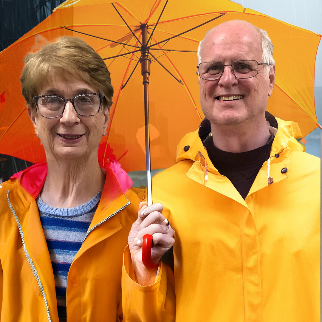 An elderly woman and a man standing close together outdoors, both smiling, holding an orange umbrella, and wearing yellow rain jackets. The woman is wearing glasses and a striped shirt underneath her rain jacket.