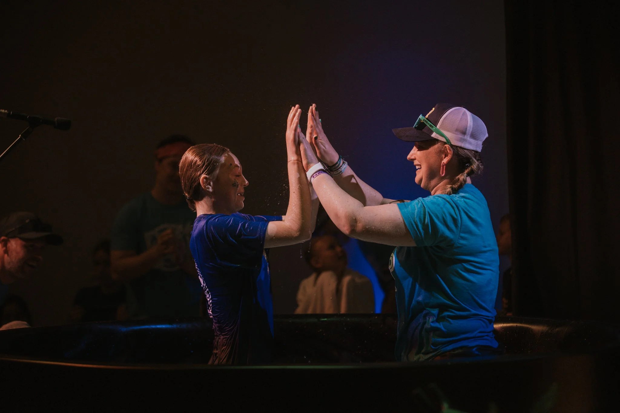 Two women in water celebrating with high-fives, one wearing a baseball cap and sunglasses, and the other with short hair and tattoos, in a dark environment with people in the background.