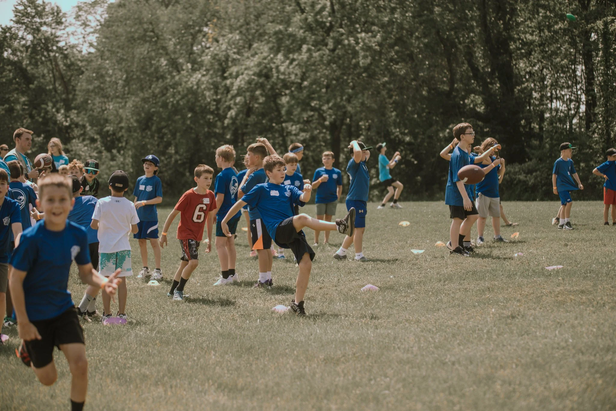 Group of boys in blue athletic clothes participating in a youth football practice or game on a grass field surrounded by trees.