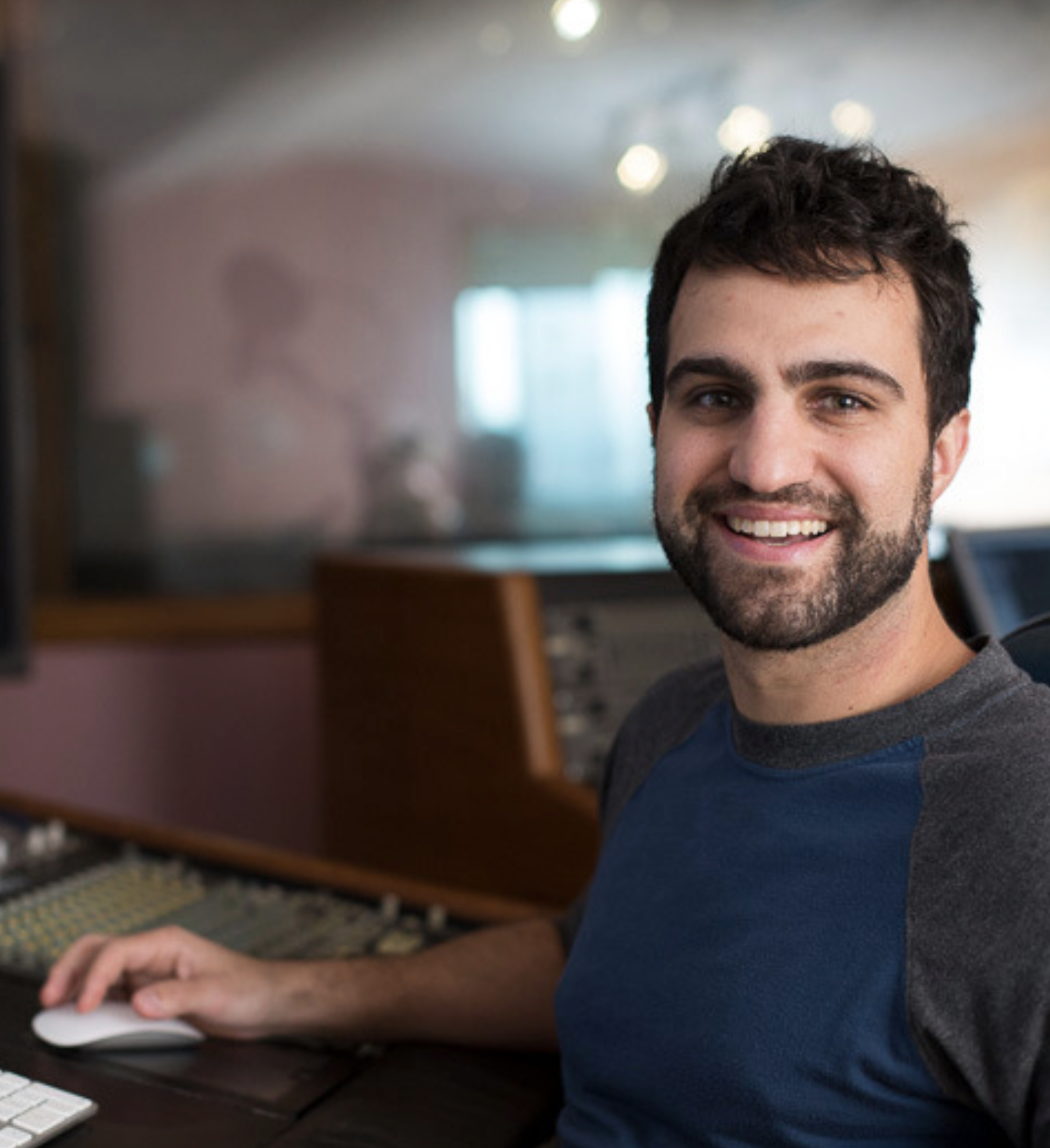 Homem sorridente em estúdio de gravação e produção musical, sentado em frente a uma mesa de áudio com equipamentos de estúdio ao fundo.