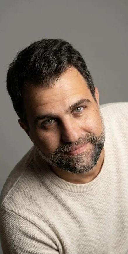 Retrato de um homem com barba e cabelo escuro, usando suéter bege claro, sorrindo levemente, contra fundo cinza.