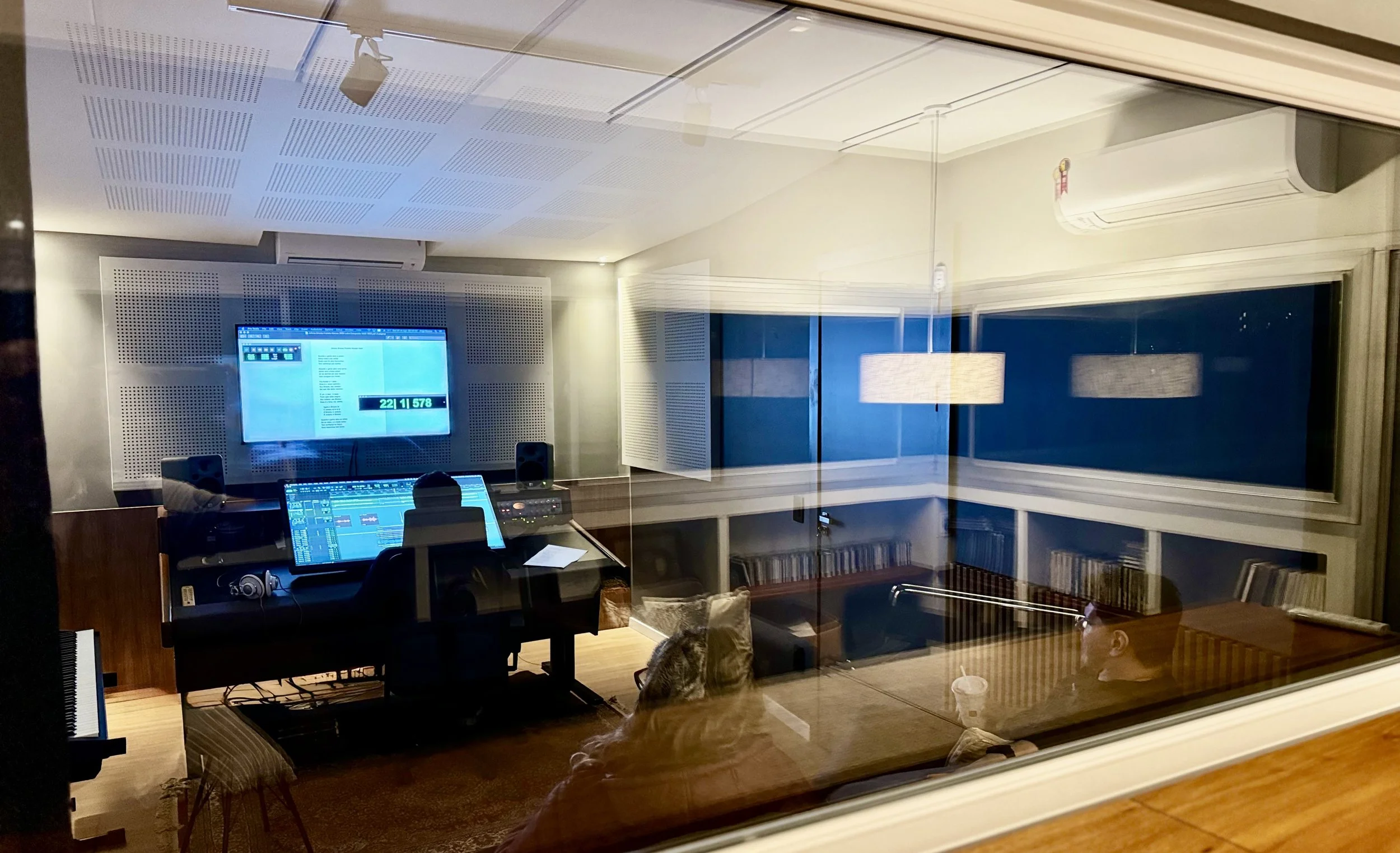 Foto de um estúdio de gravação de música moderno. Entre reflexos de janelas, a mesa de gravação é uma tela sensível ao toque. Um téncnico trabalha, duas pessoas assistem.