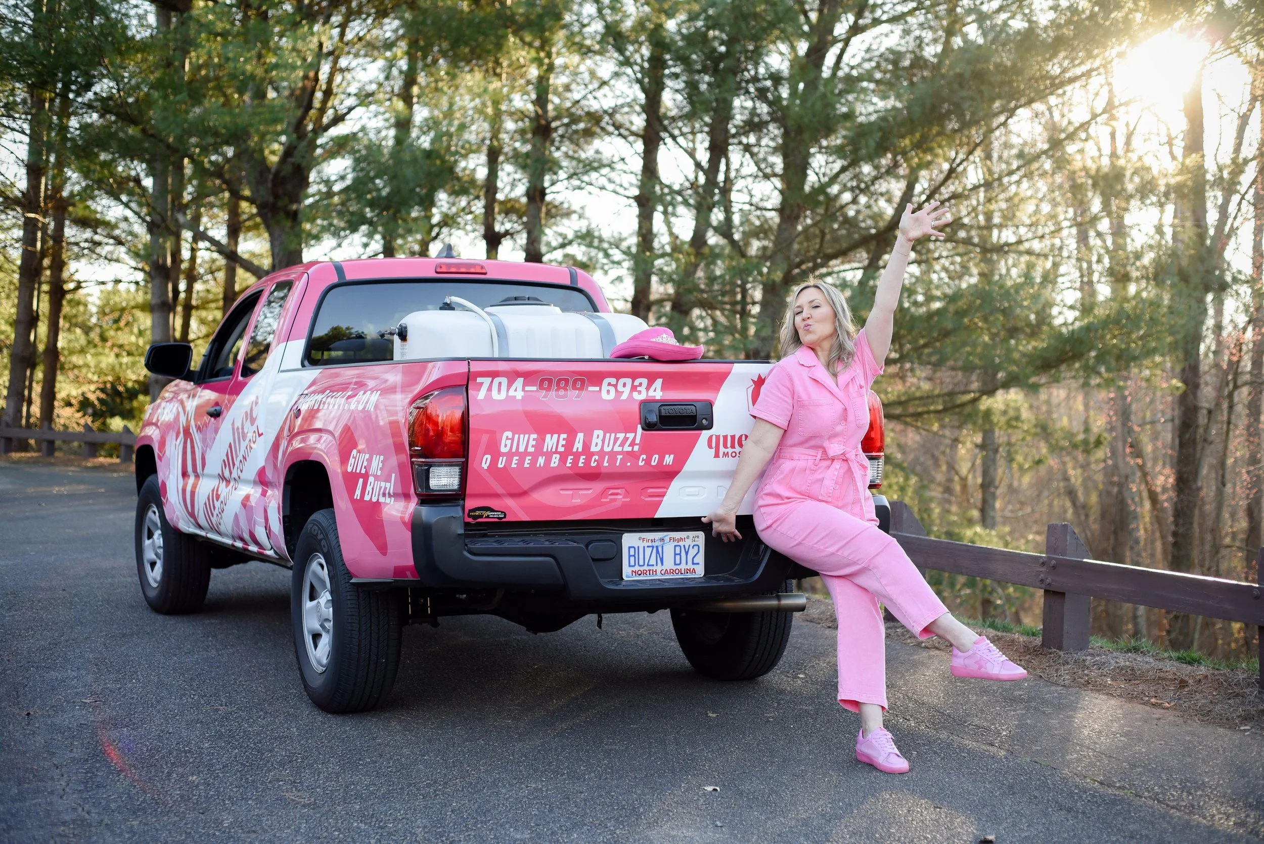 A woman dressed in pink jumpsuit and pink sneakers leaning against the back of a pink pickup truck with water tanks and pink accessories, posing with one arm raised and blowing a kiss, in a wooded area during sunset.