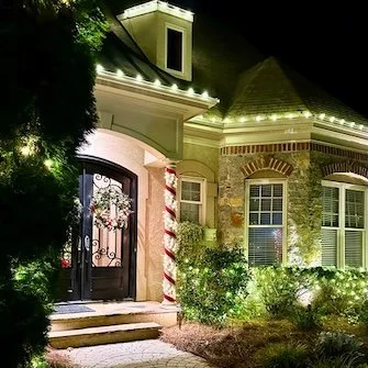 Night view of a house decorated with Christmas lights, a wreath on the door, and candy cane-striped columns, with bushes and trees illuminated in the yard.