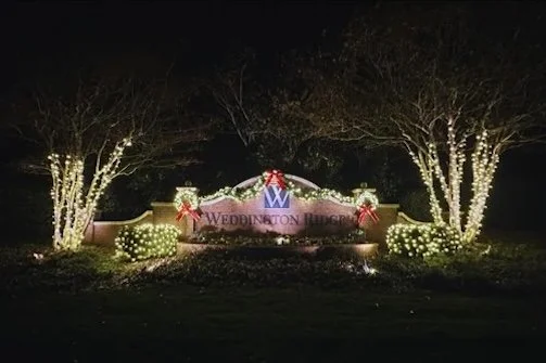 Decorative sign for Weddington Ridge, illuminated with Christmas lights, wreaths, and red bows at night.