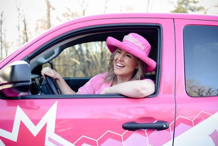 A woman in a pink dress and large pink hat with a lace band, smiling and sitting in the driver's seat of a pink car with a geometric white and pink pattern. She is outdoors with trees in the background.