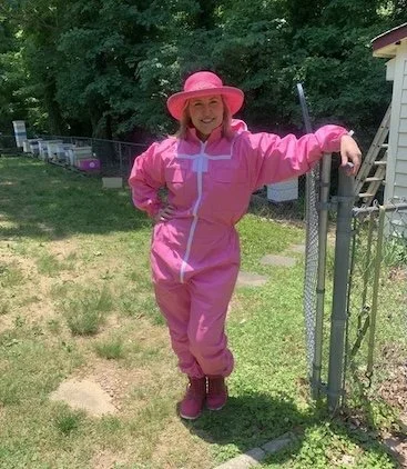 A woman wearing a pink hat, pink jacket, and pink boots standing outdoors next to a chain-link fence with a garden or yard in the background.