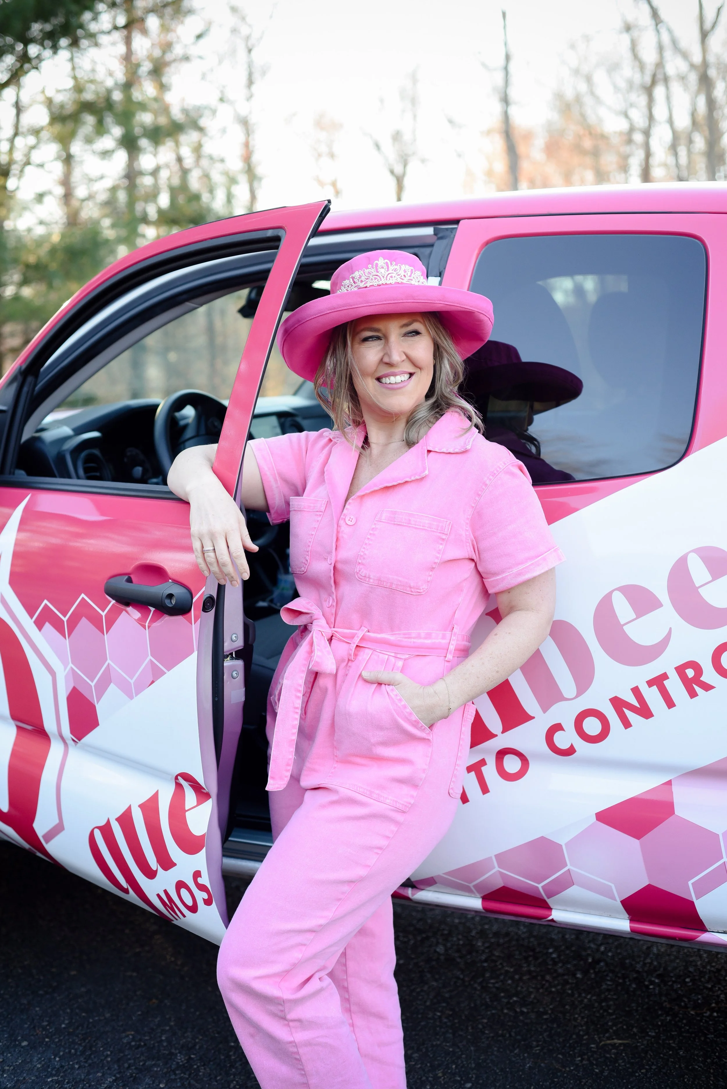 Woman in pink jumpsuit and large pink hat standing outside a pink campaign car with pink and white honeycomb pattern, smiling.
