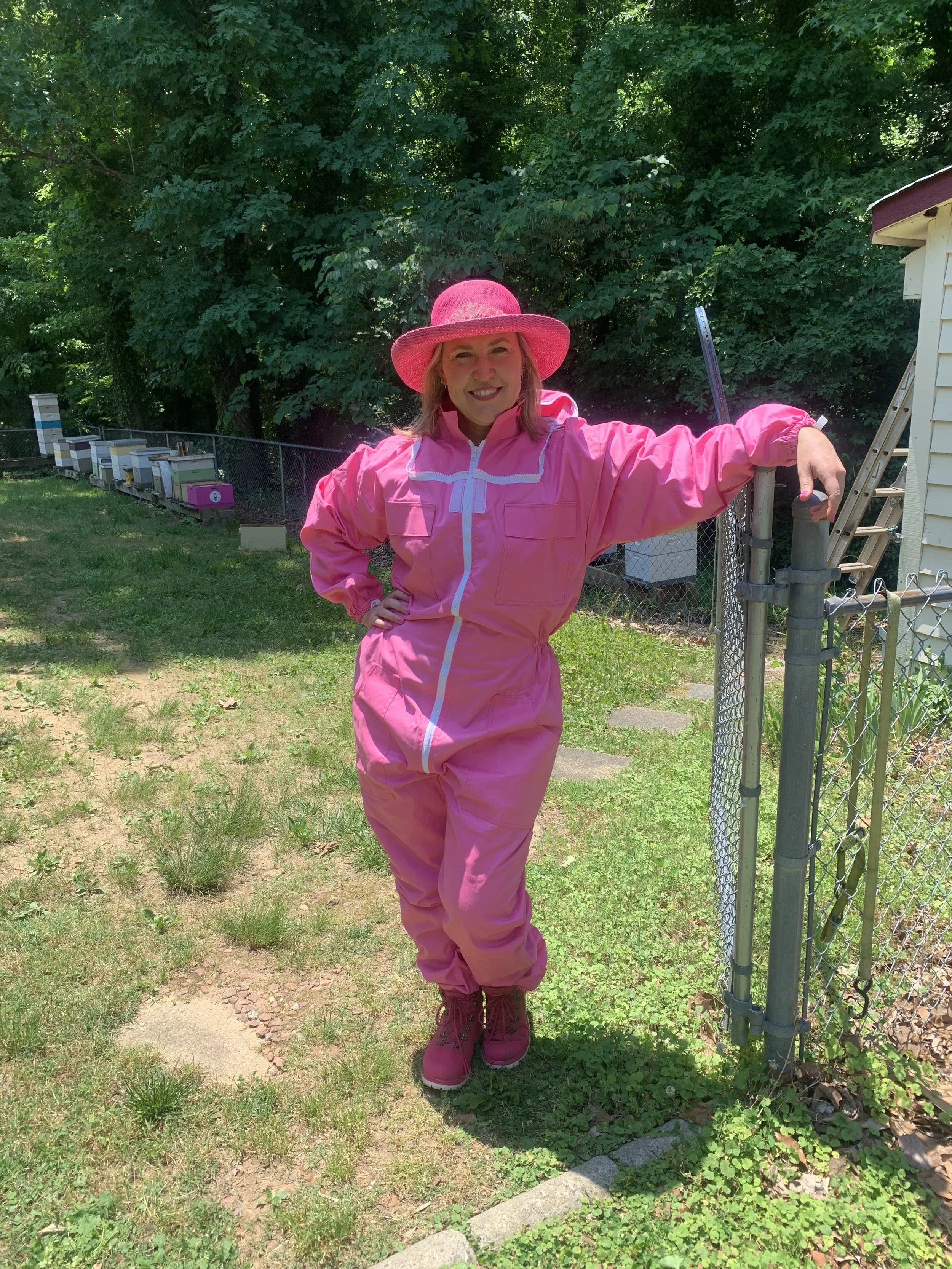 A woman wearing a pink hat, pink jacket, and pink boots standing outdoors next to a chain-link fence with a garden or yard in the background.