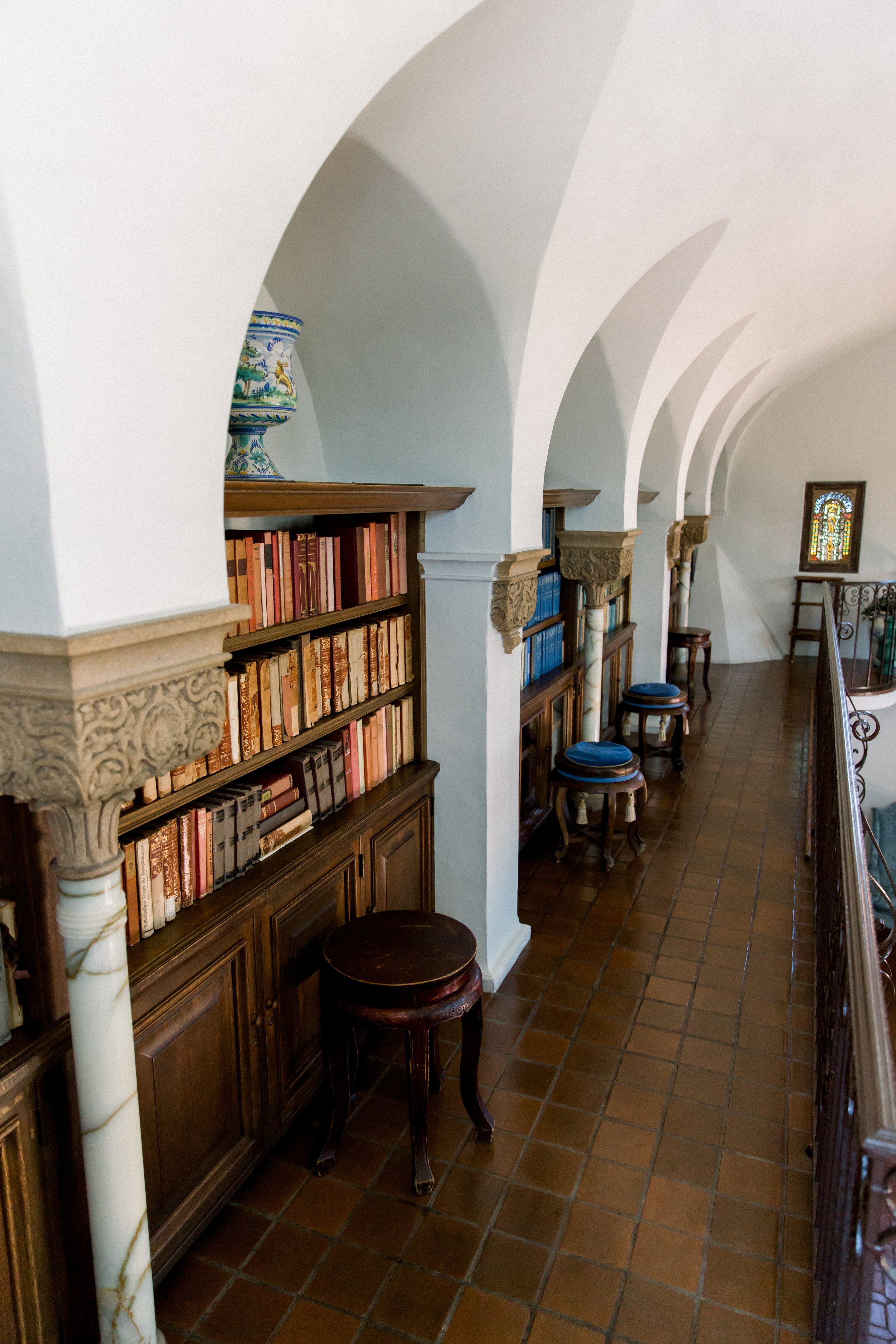 Library hallway with arched ceiling, bookshelves, stools, and decorative vase.