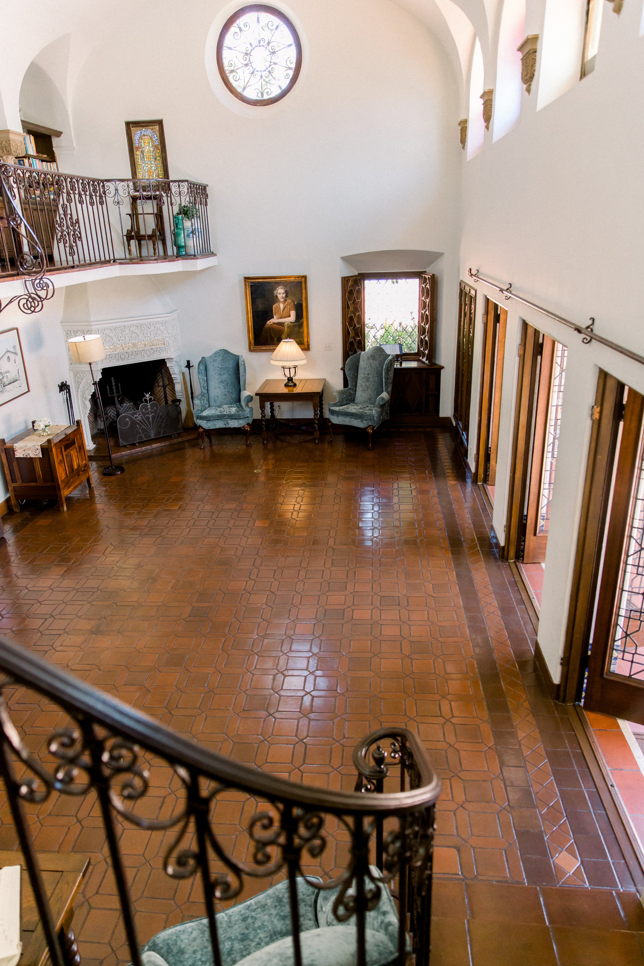 Cozy living room with high ceiling, wood tile floor, and elegant staircase. Features two blue armchairs, a painting, a fireplace, a large window, and an upstairs railing with decorative metalwork.