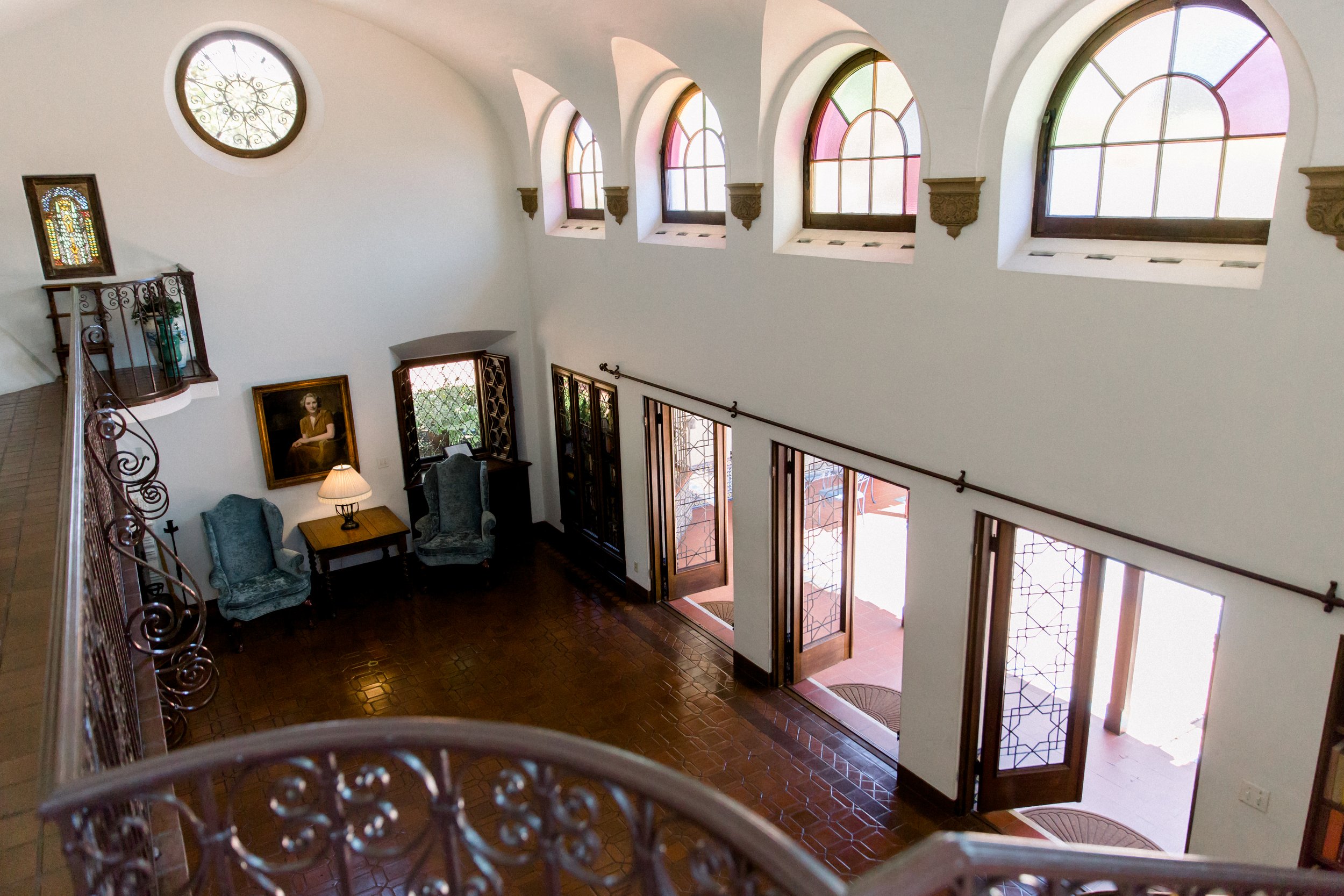 Elegant room interior with arched windows and iron railings, showcasing a sitting area with two armchairs, a portrait painting, and glass-paneled doors leading outside.