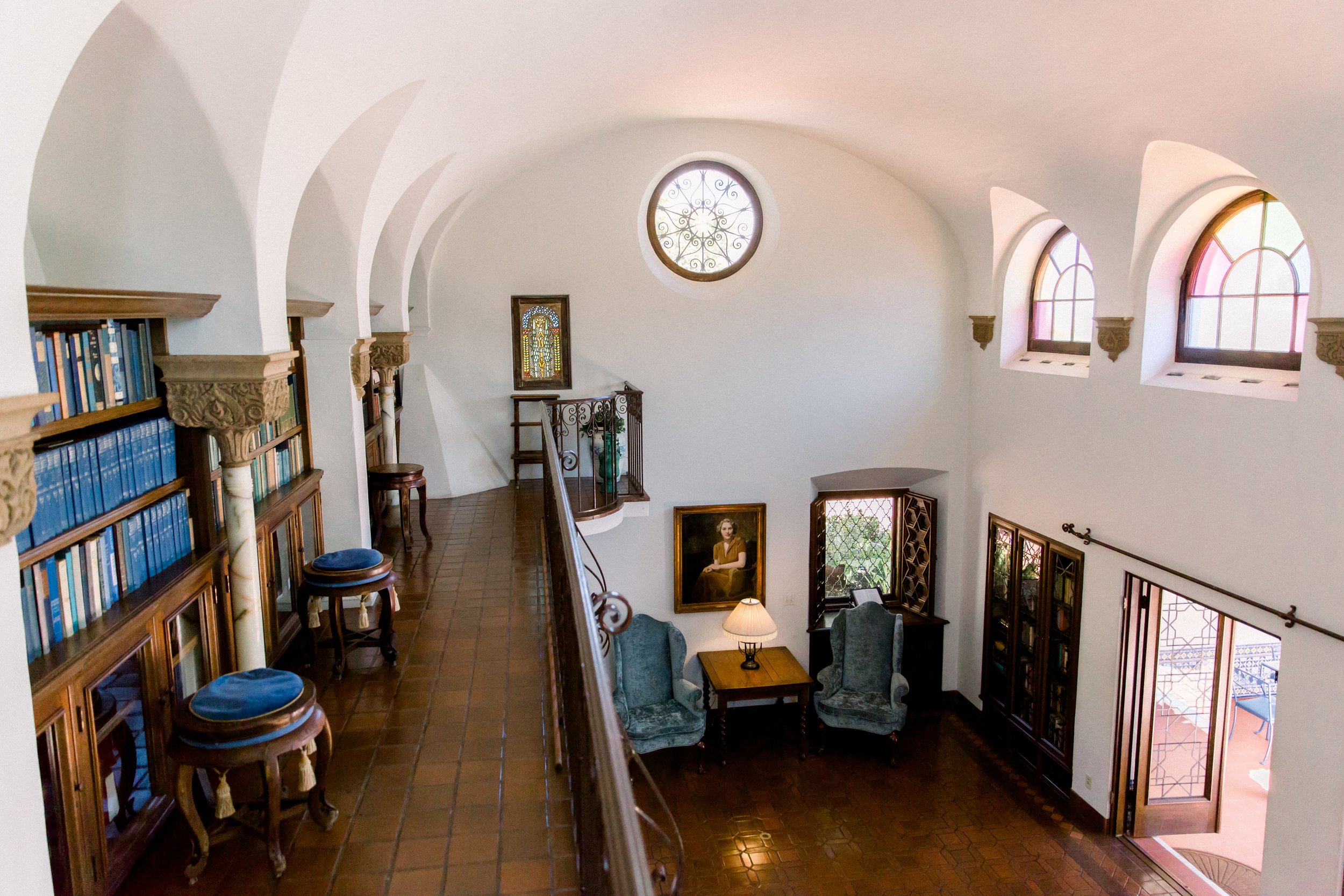 Elegant two-story study with arched windows, bookshelves, and a lounge area with armchairs and a table with a lamp.