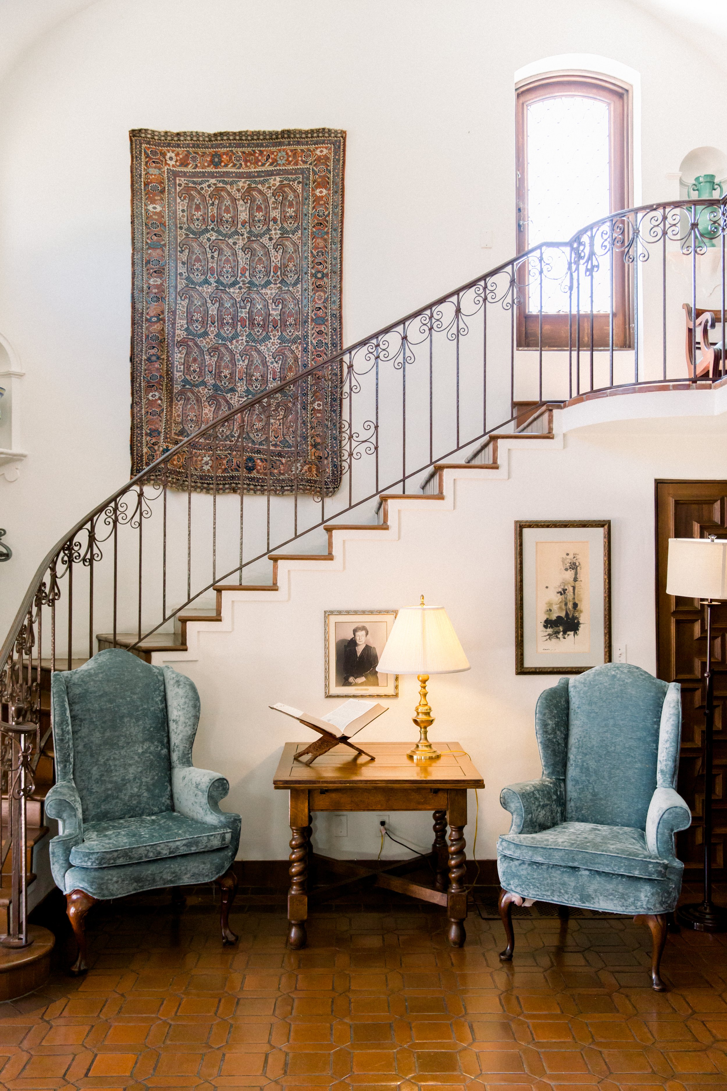 Cozy living area with two blue vintage armchairs, a wooden side table, a lamp, and framed pictures. A decorative carpet hangs on a staircase with ornate railing.