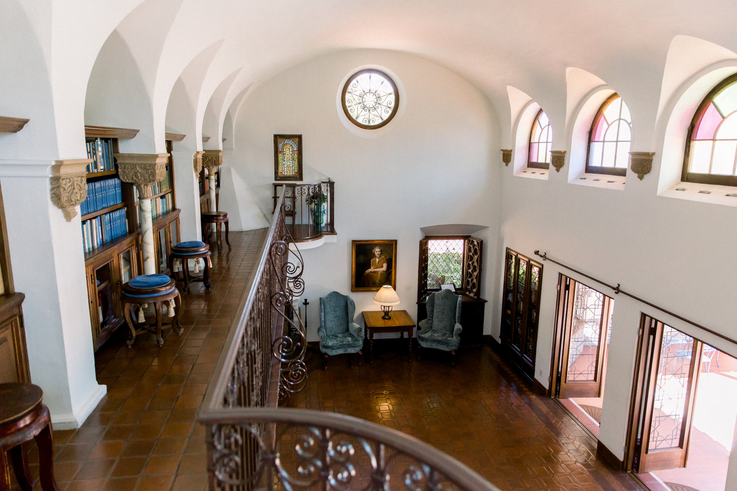 Interior of a Spanish-style room with arched doorways, colorful stained glass windows, wooden bookshelves, and a wrought iron railing. The room features vintage furniture, including blue upholstered chairs and a wooden side table with a lamp. A large