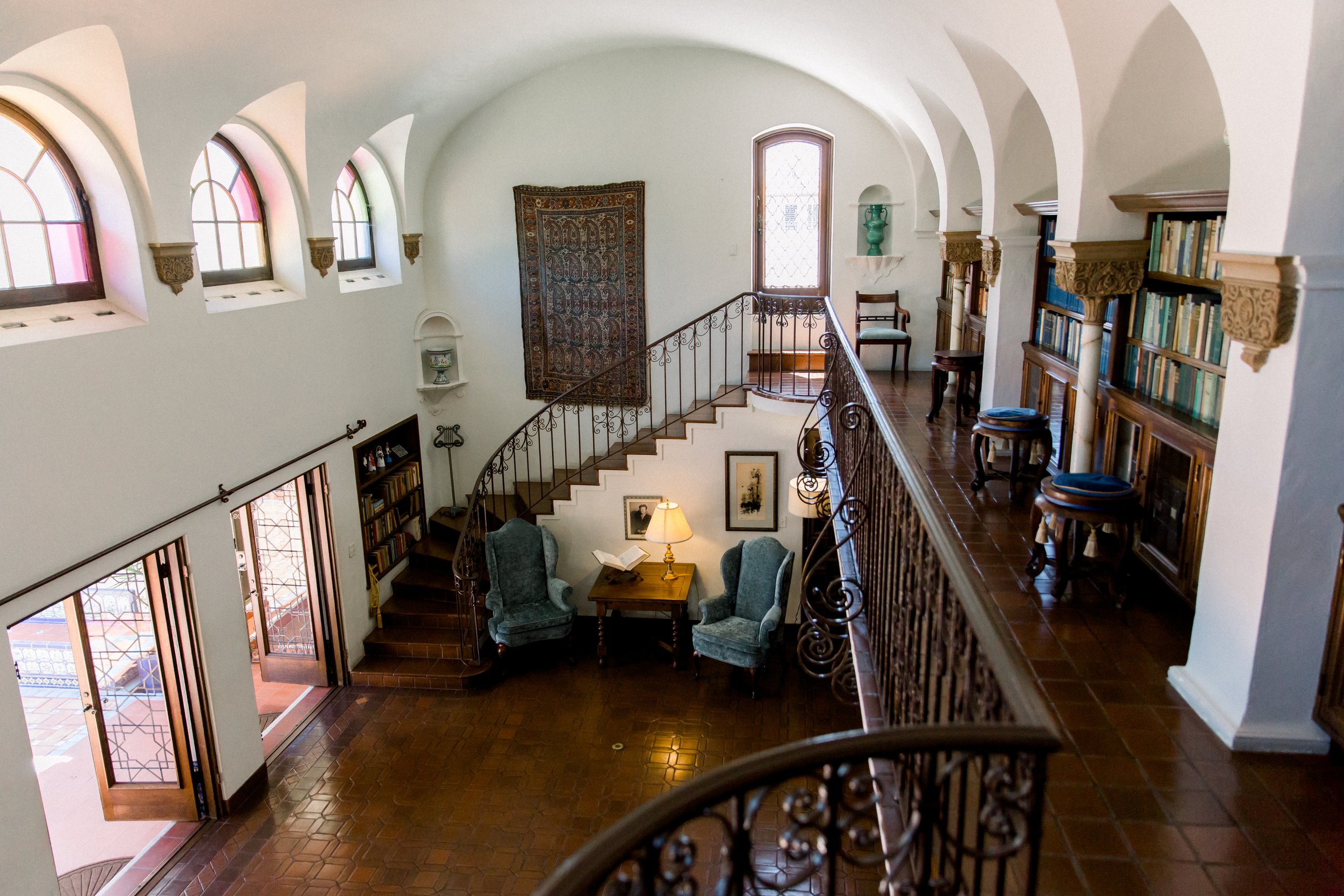 Elegant interior with arched windows, a curved staircase, bookshelves, and two armchairs around a small table with a lamp.