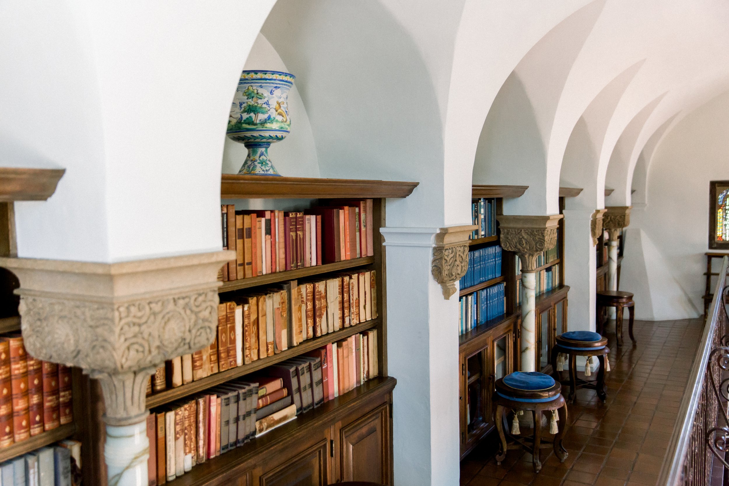 Bookshelves in a library with decorative arches and a ceramic vase, featuring books with colorful spines and wooden stools.