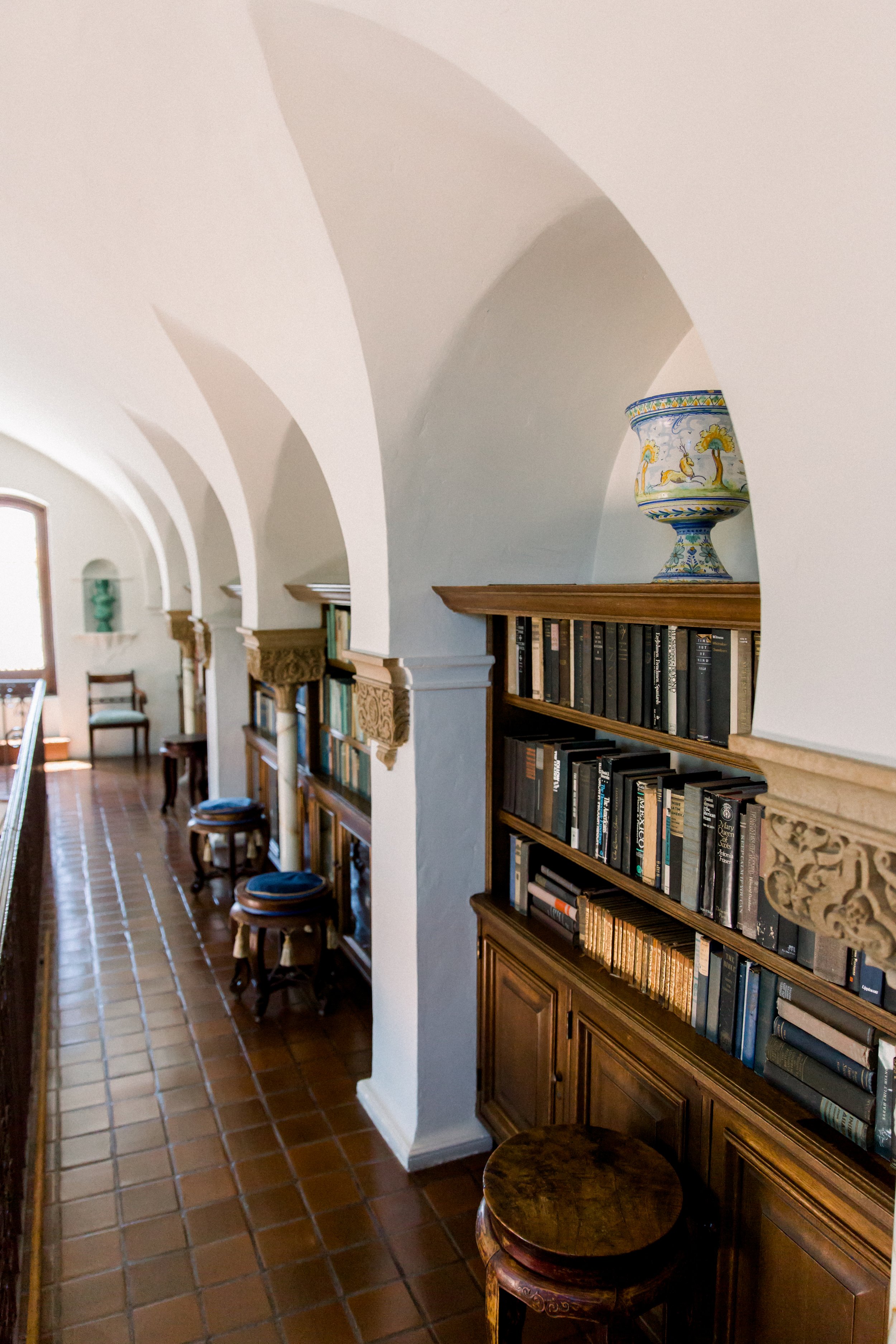 Library with wooden bookshelves, vaulted ceiling, decorative columns, stools, and a large decorative vase on top shelf.