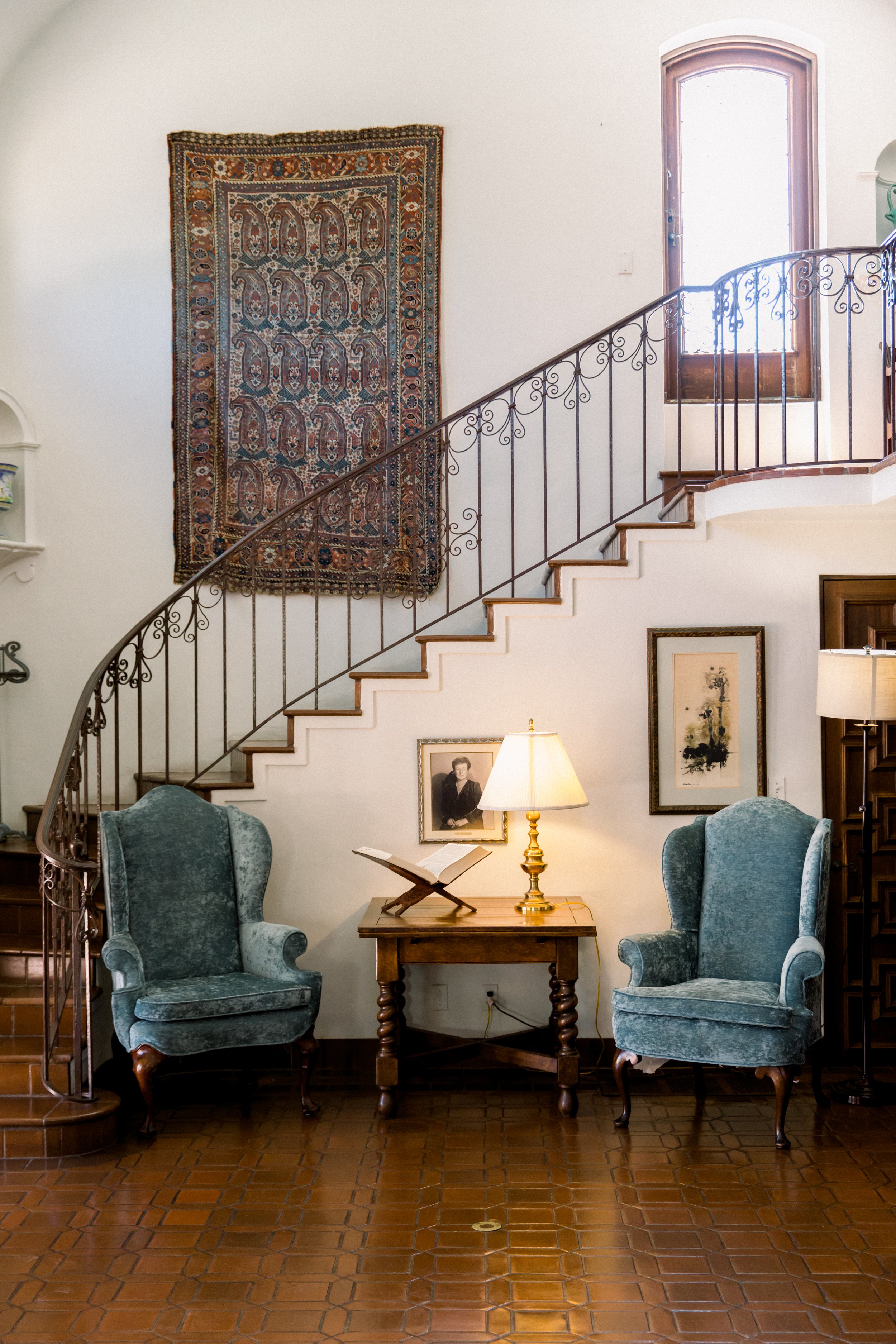 An elegant living room featuring a curved staircase with an ornate railing, a decorative woven wall hanging, two blue velvet armchairs, a wooden side table with a lamp, and framed artwork on the wall.