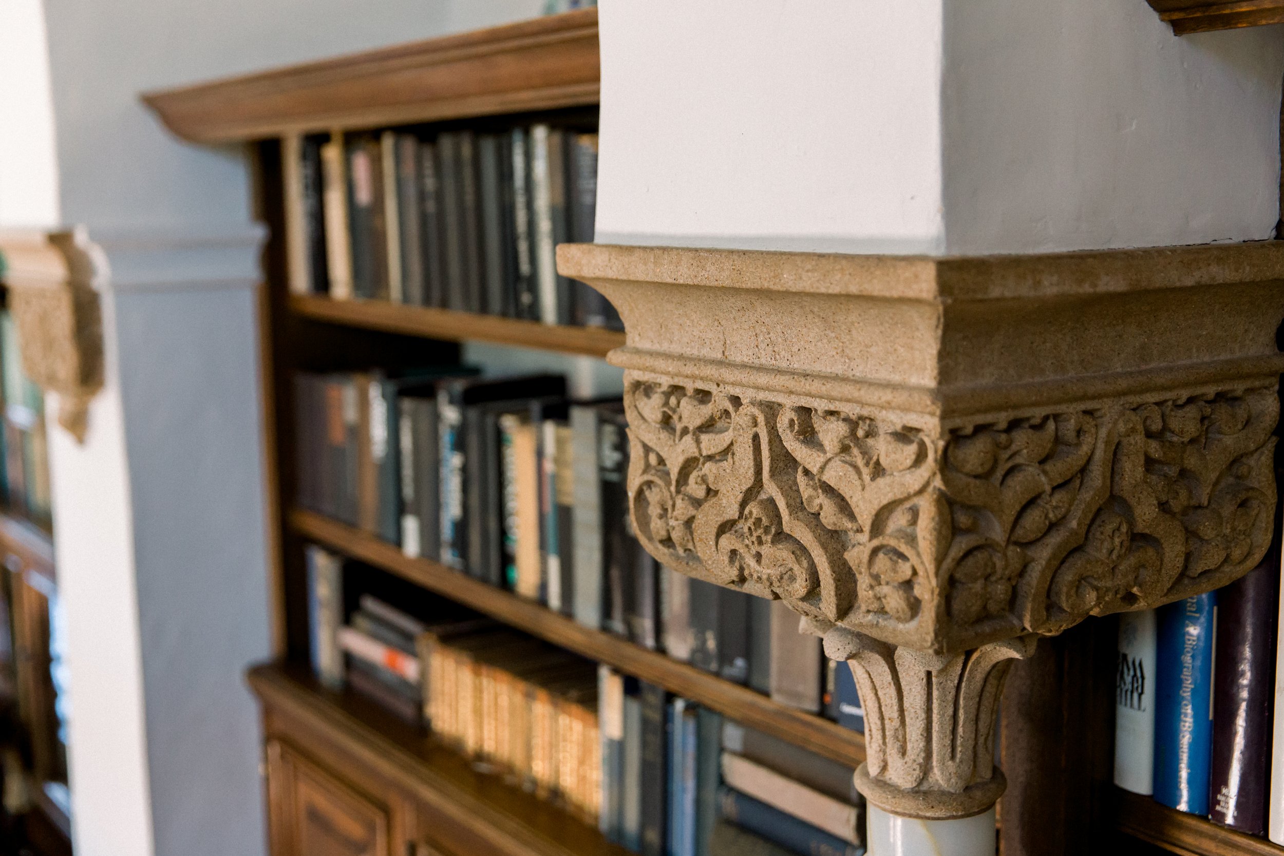 Ornate stone shelf bracket with floral patterns in a library setting, with bookshelves in the background.