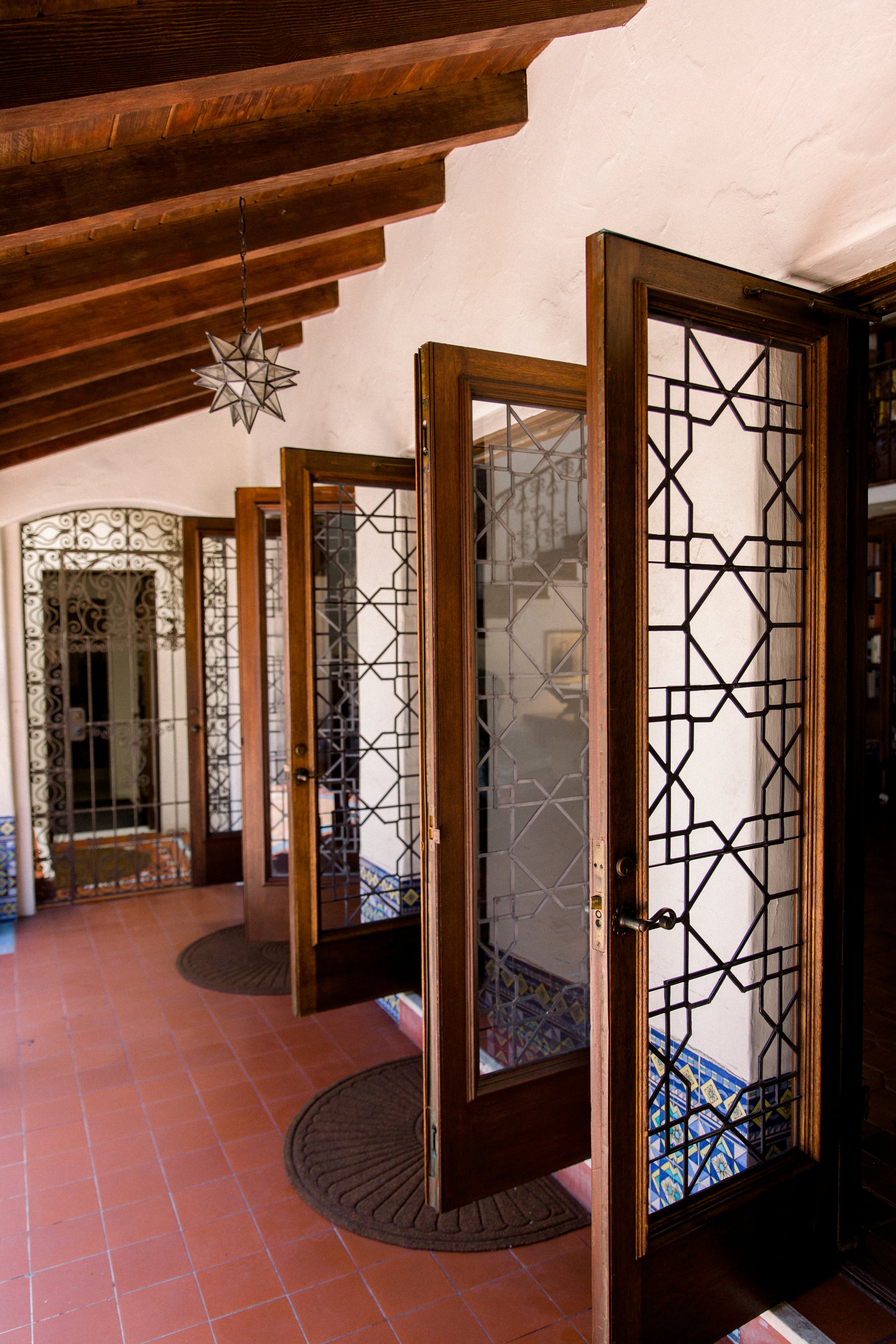 Spanish-style hallway with wooden doors featuring decorative glass panels, red tile flooring, blue patterned tiles, and a star-shaped light fixture.