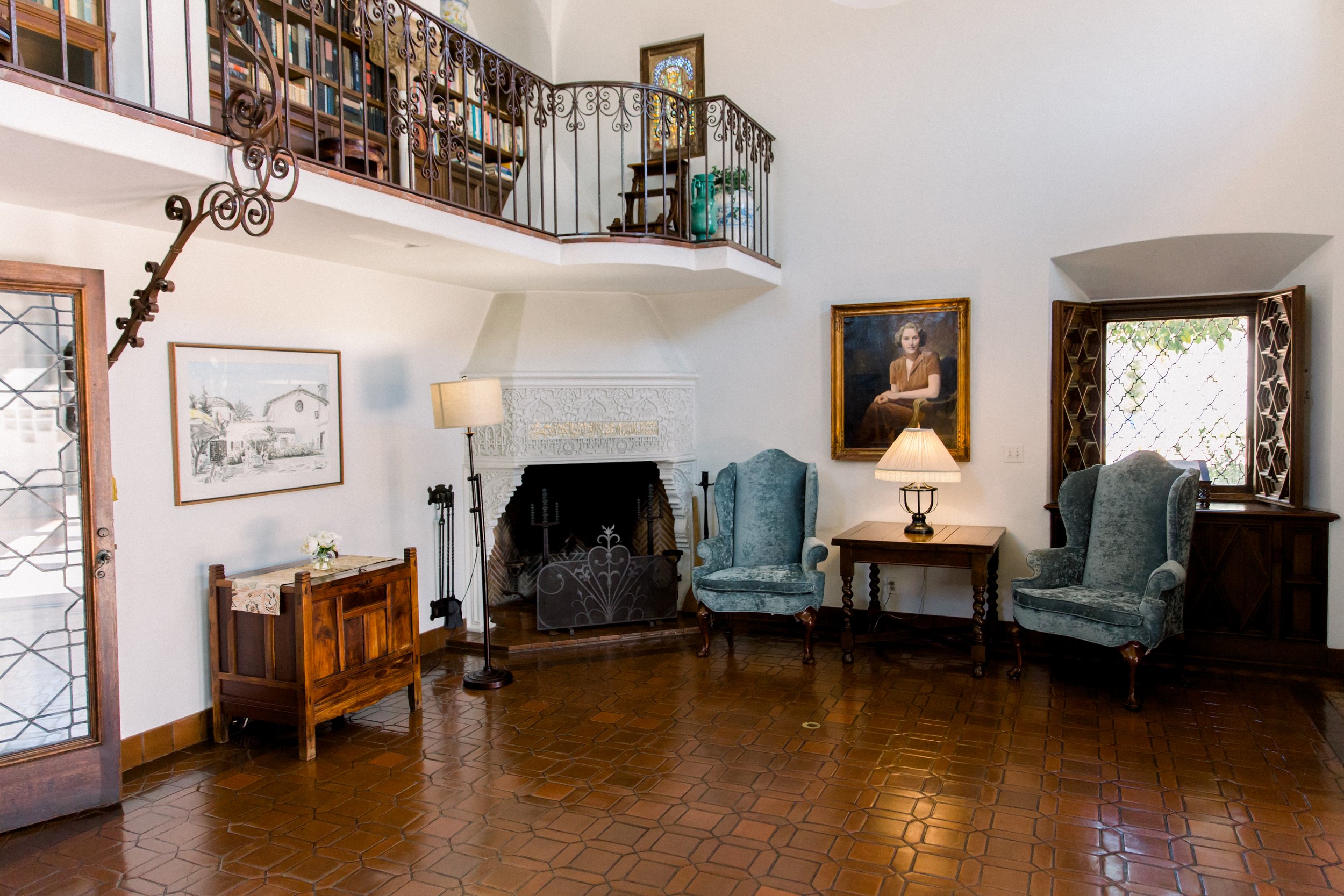 Elegant living room with a high ceiling, featuring a fireplace, two upholstered chairs, a table with a lamp, and a staircase leading to an upper level with a bookcase. A portrait hangs on the wall, and the floor is tiled.
