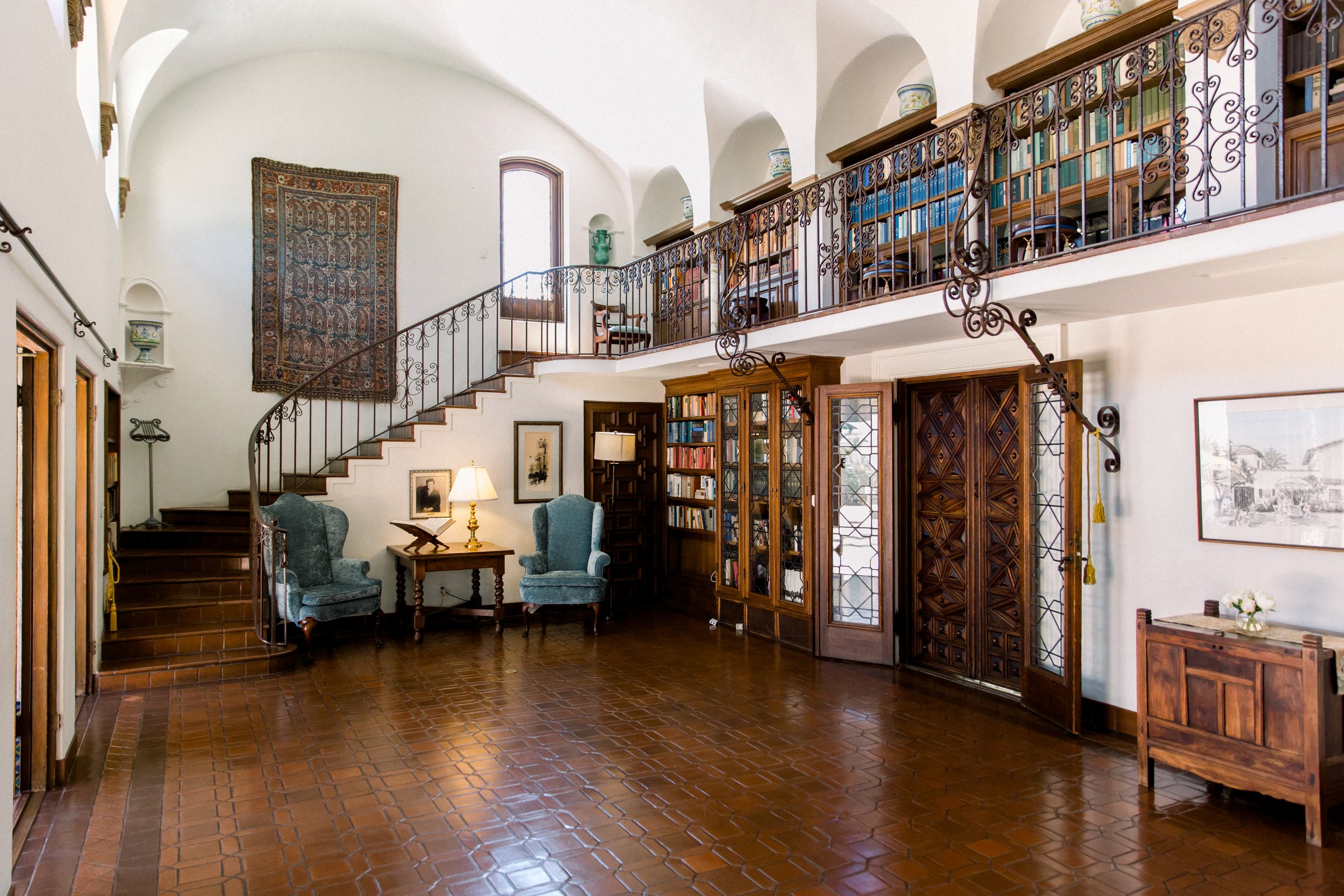 Elegant living room with large bookcase, armchairs, spiral staircase, and decorative rug on wall.