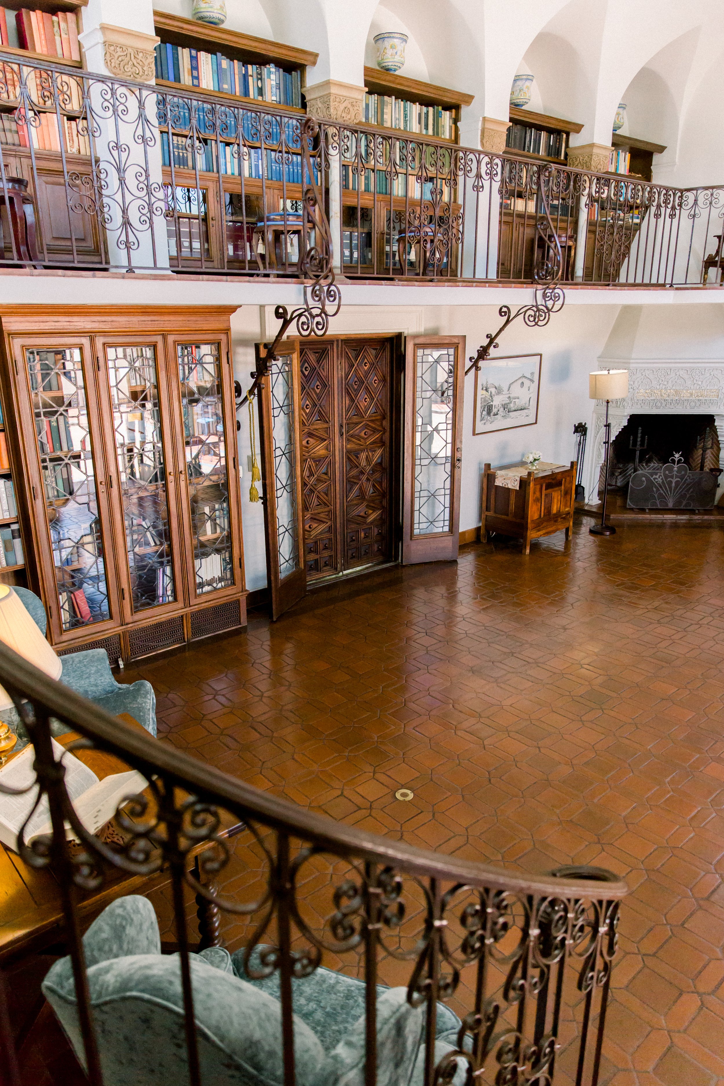Interior of a vintage library with a mezzanine, ornate metal railings, bookshelves, wooden furniture, and a tiled floor. The room includes a fireplace, a decorative lamp, and a seating area.