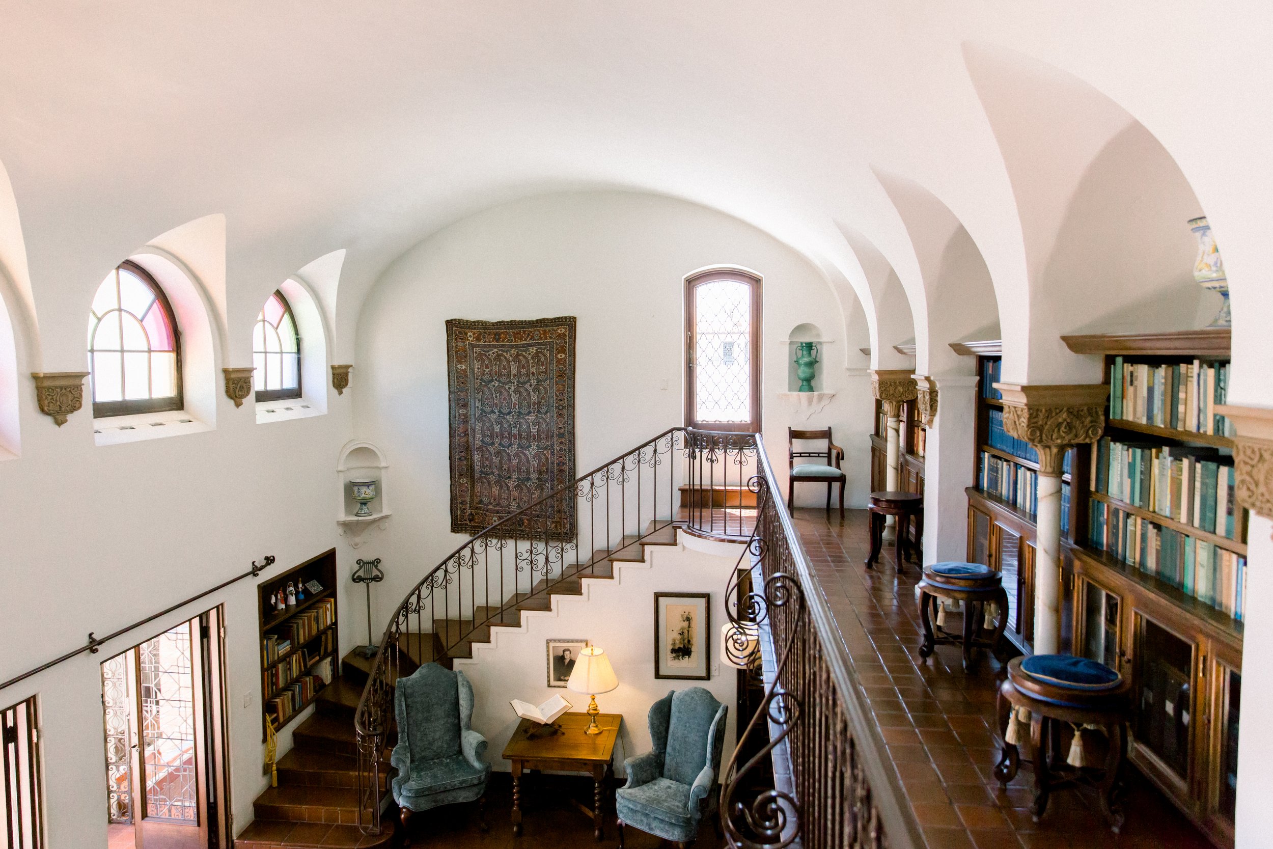 Interior of a library with arched windows, ornate railing, bookshelves, armchairs, and a wall tapestry.