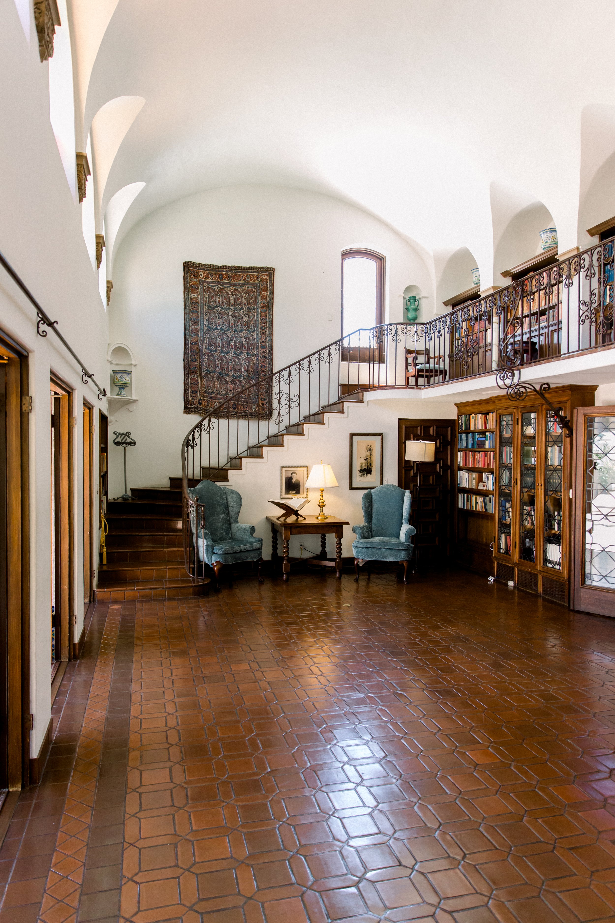 Elegant interior of a spacious room featuring a curving staircase with wrought iron railing, blue upholstered chairs, a wooden side table with a lamp, bookshelves filled with books, and a large decorative rug mounted on the wall. The room has high ce