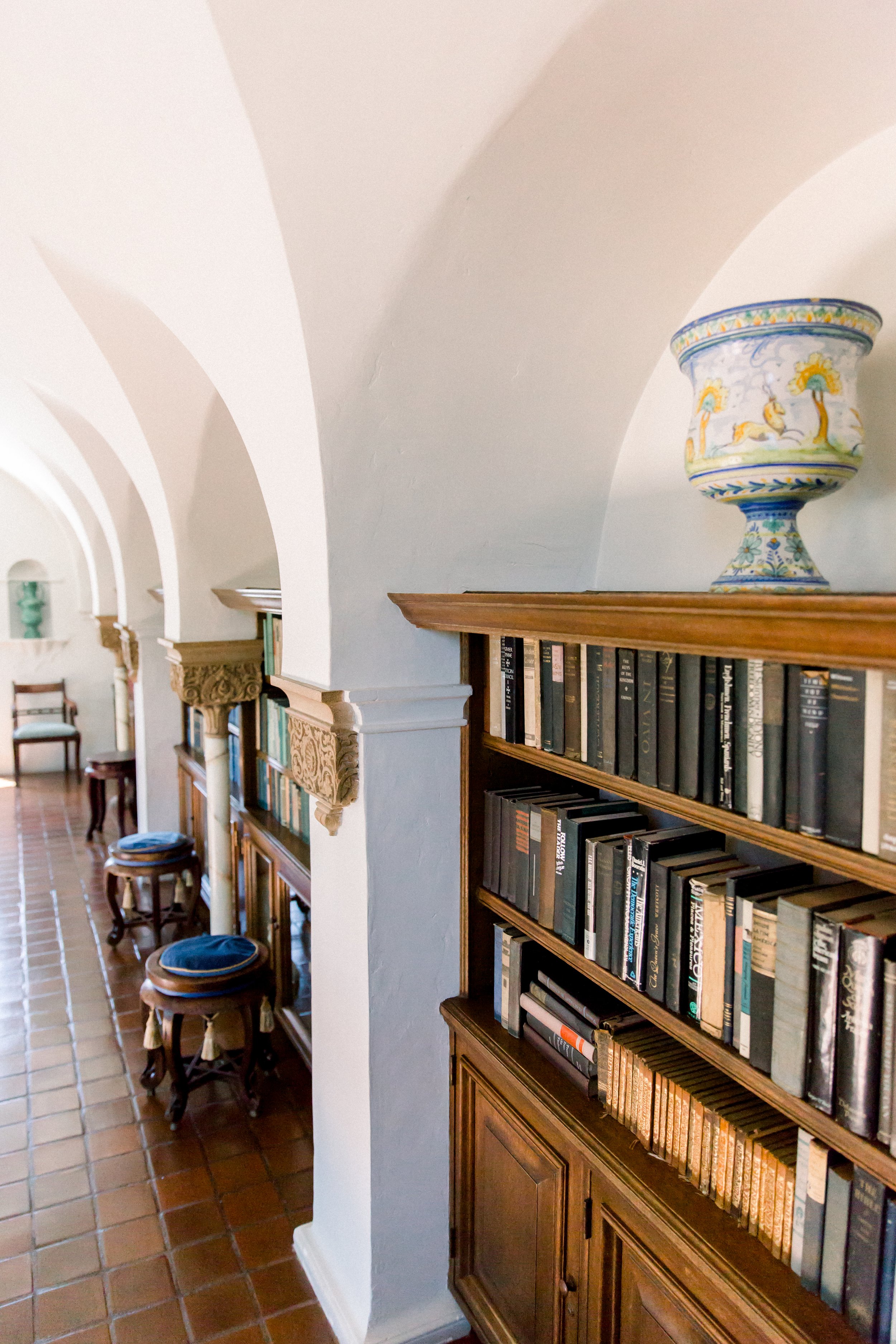 A hallway with tiled floor, arched ceilings, bookshelves filled with books, decorative corbels, stools with blue cushions, and a large ornamental vase on top of the bookshelf.