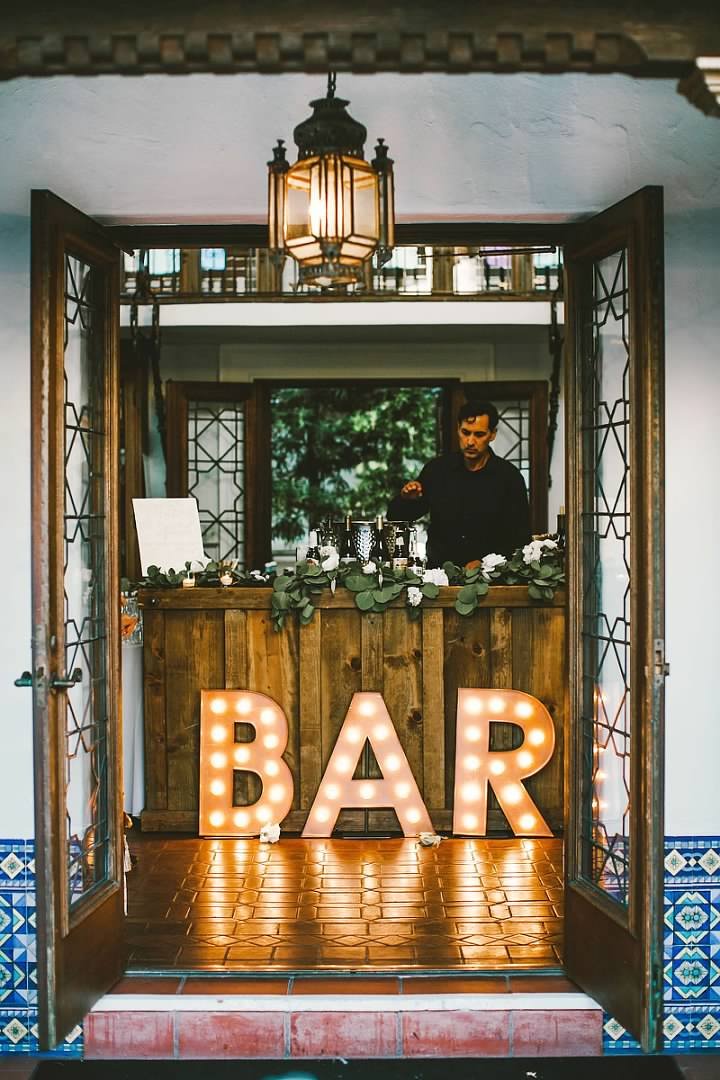 Outdoor bar with illuminated "BAR" sign and a bartender, decorated with flowers and plants.