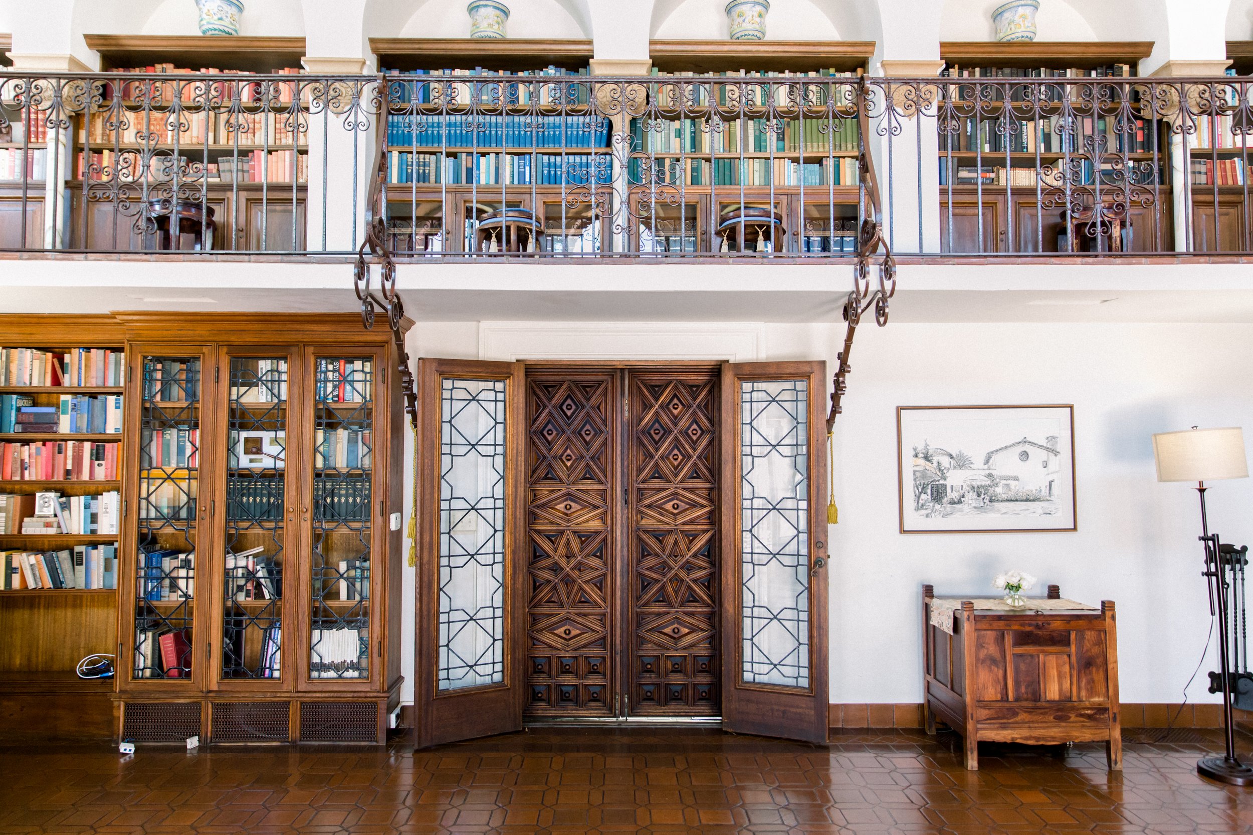 Elegant library with wooden bookshelves, decorative railing, wooden double doors, and framed artwork on the wall.