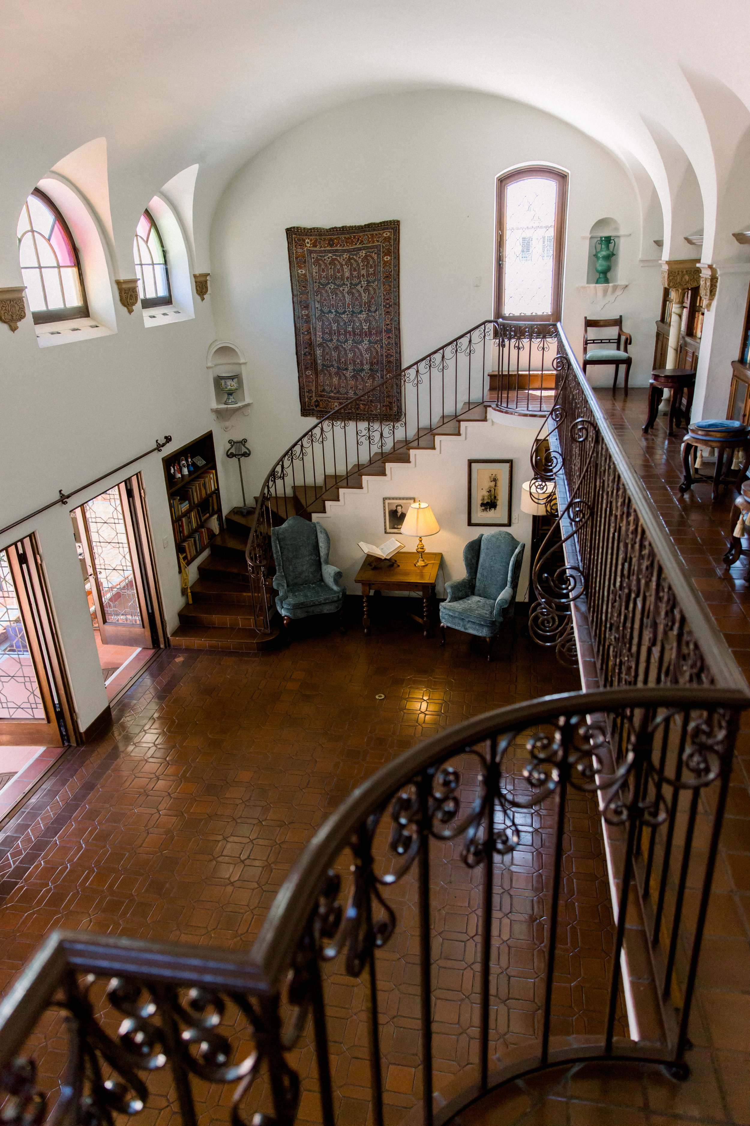 Interior of a room with a curved staircase, ornate iron railings, two blue armchairs, a wooden table with a lamp, and arched windows on a white wall.