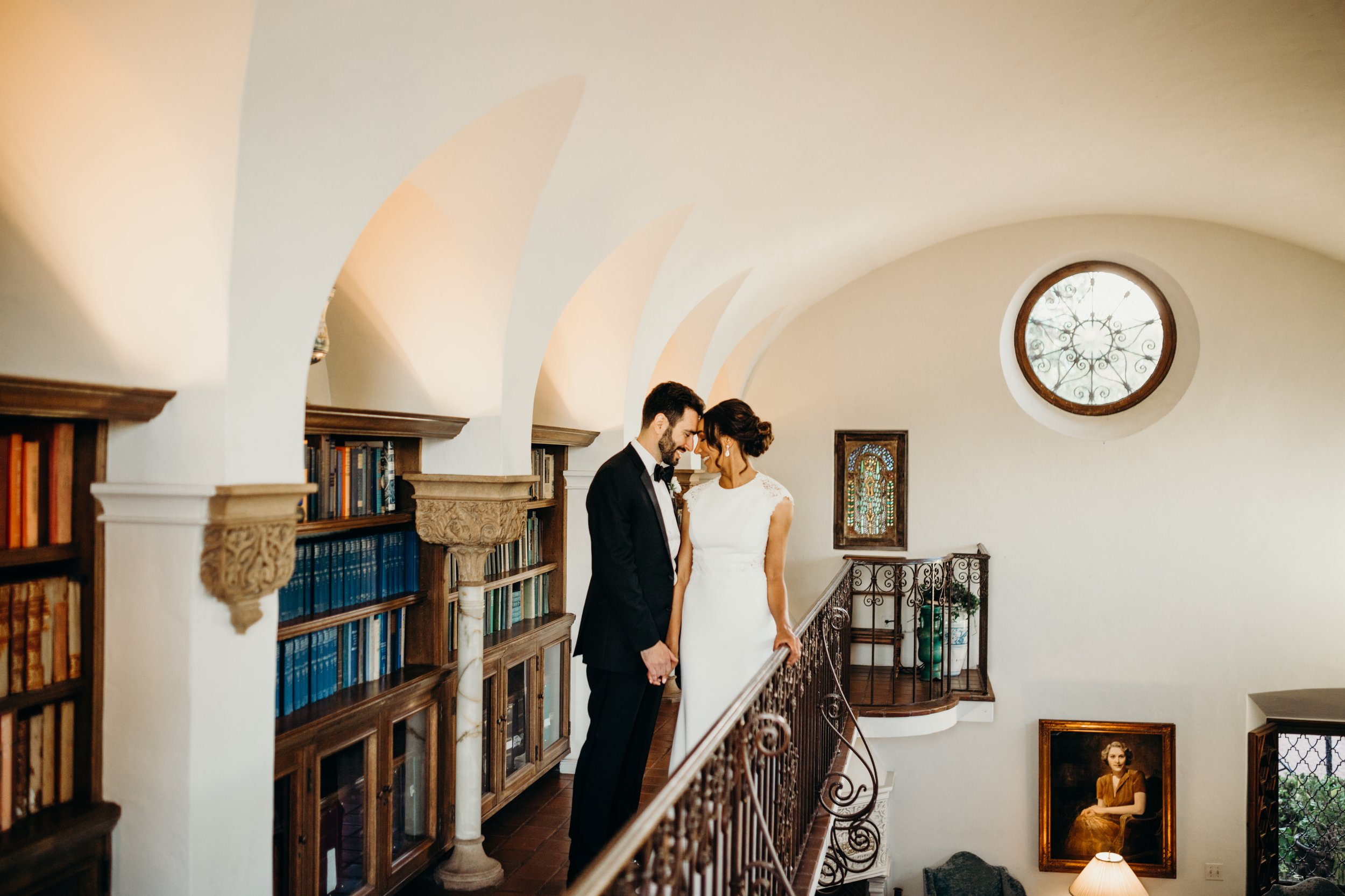 Elegant couple standing in a library next to bookshelves and ornate railings, with a round window above and a portrait on the wall.