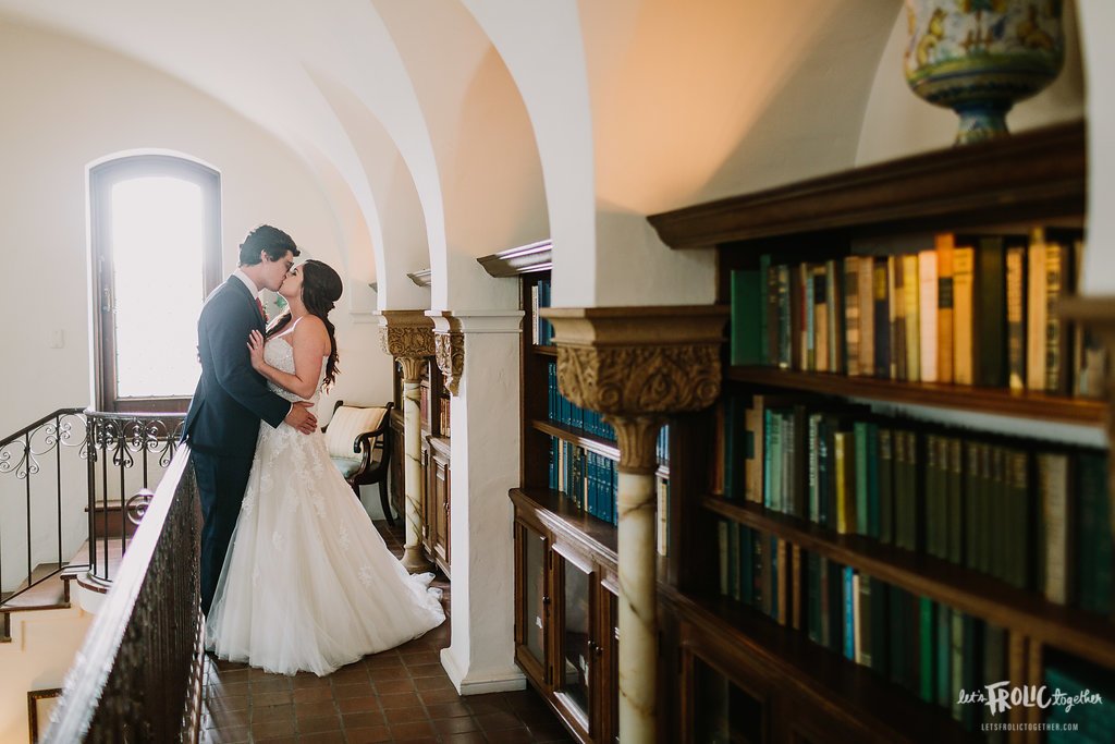 A bride and groom kiss in an elegant library with arched ceilings, surrounded by bookshelves filled with books. The bride wears a white dress, and the groom a dark suit.