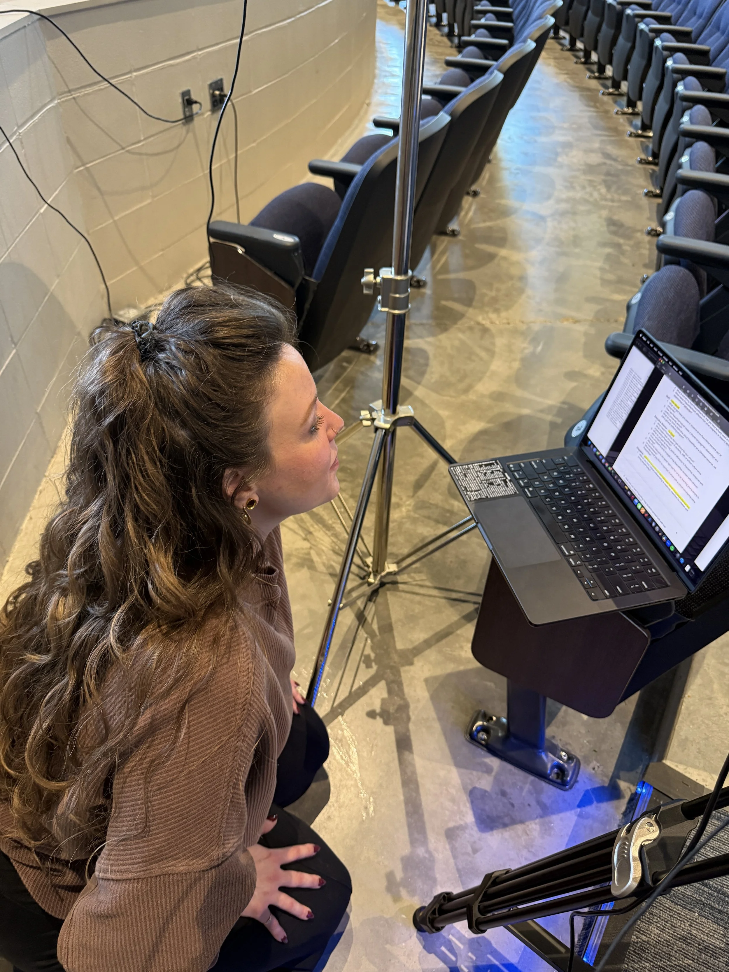 A woman with curly brown hair, earrings, and a brown jacket sitting at a table with a laptop, in an auditorium or conference room with rows of blue padded chairs.