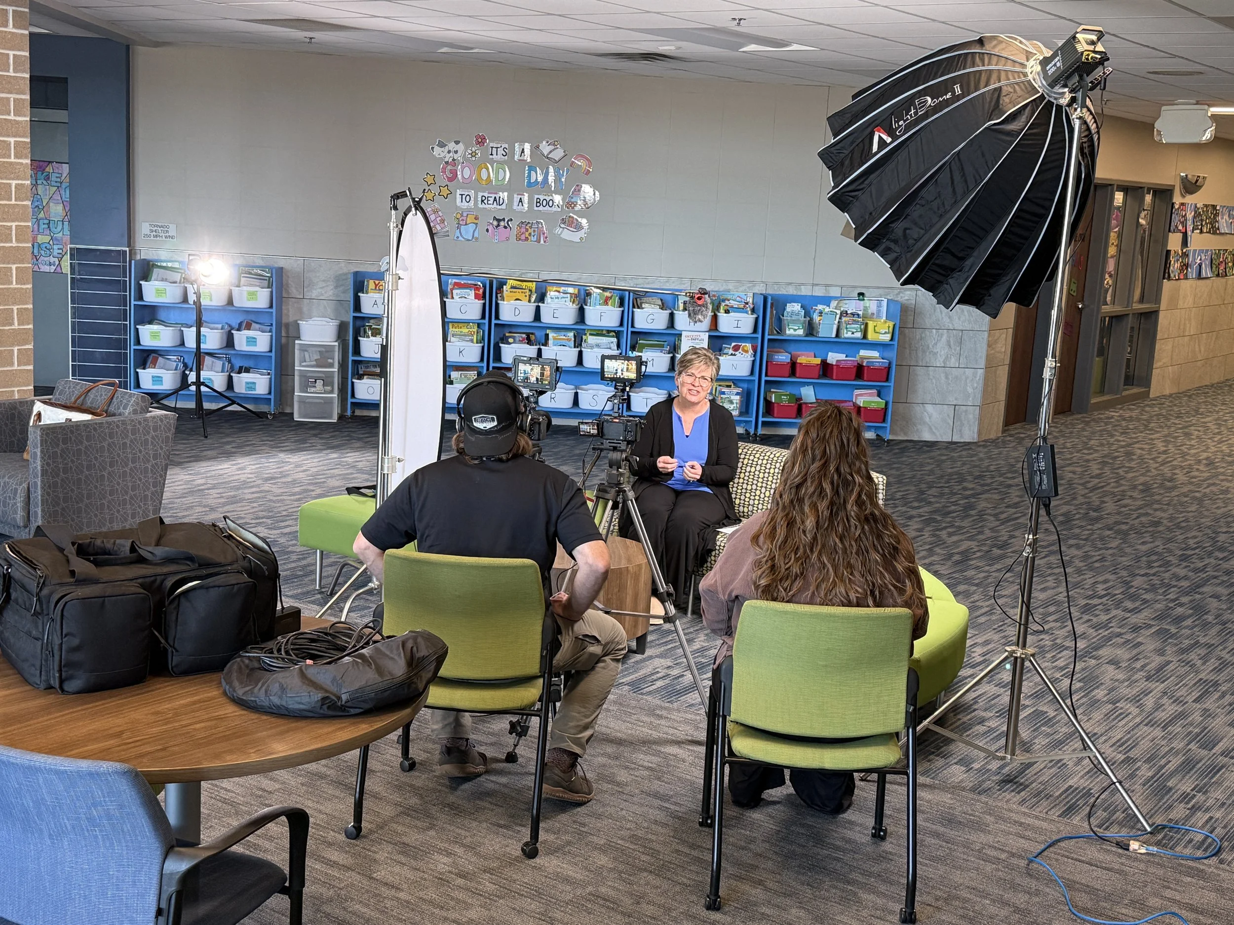 Indoor scene with a woman speaking to people in a small event setup. The setup includes lighting equipment, cameras, and chairs, with bookshelves and colorful signs on the wall reading 'It's a good day to read a book' in the background.