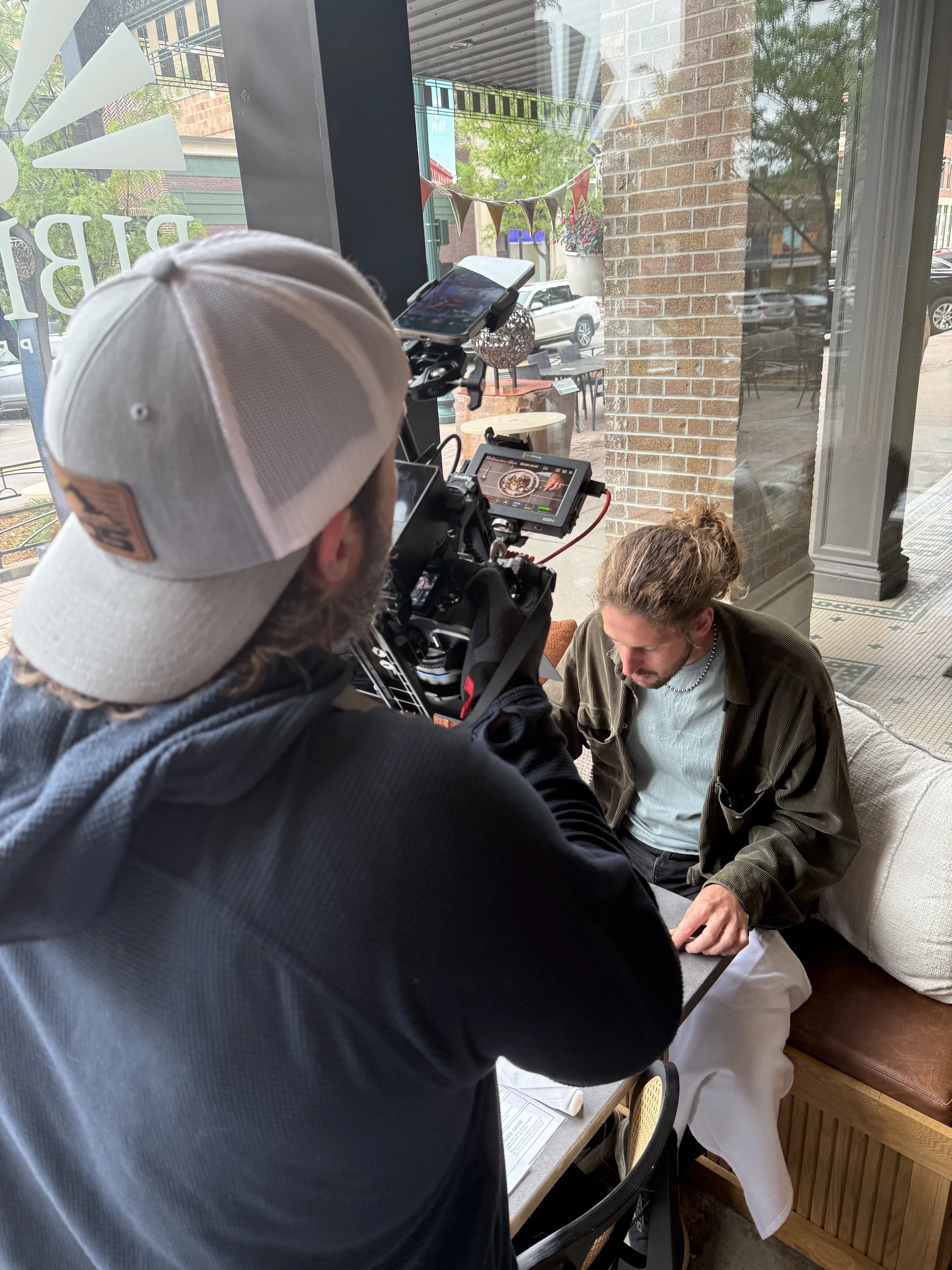 A man wearing a beige baseball cap and a dark jacket uses a professional camera to shoot a photo inside a café. He is capturing a woman sitting at a table, dressed in a green jacket and white pants, who is engaged in the photo.