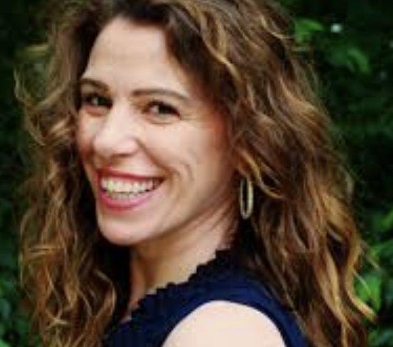 Smiling woman with curly brown hair in a dark blue top, outdoors, with green foliage in the background.