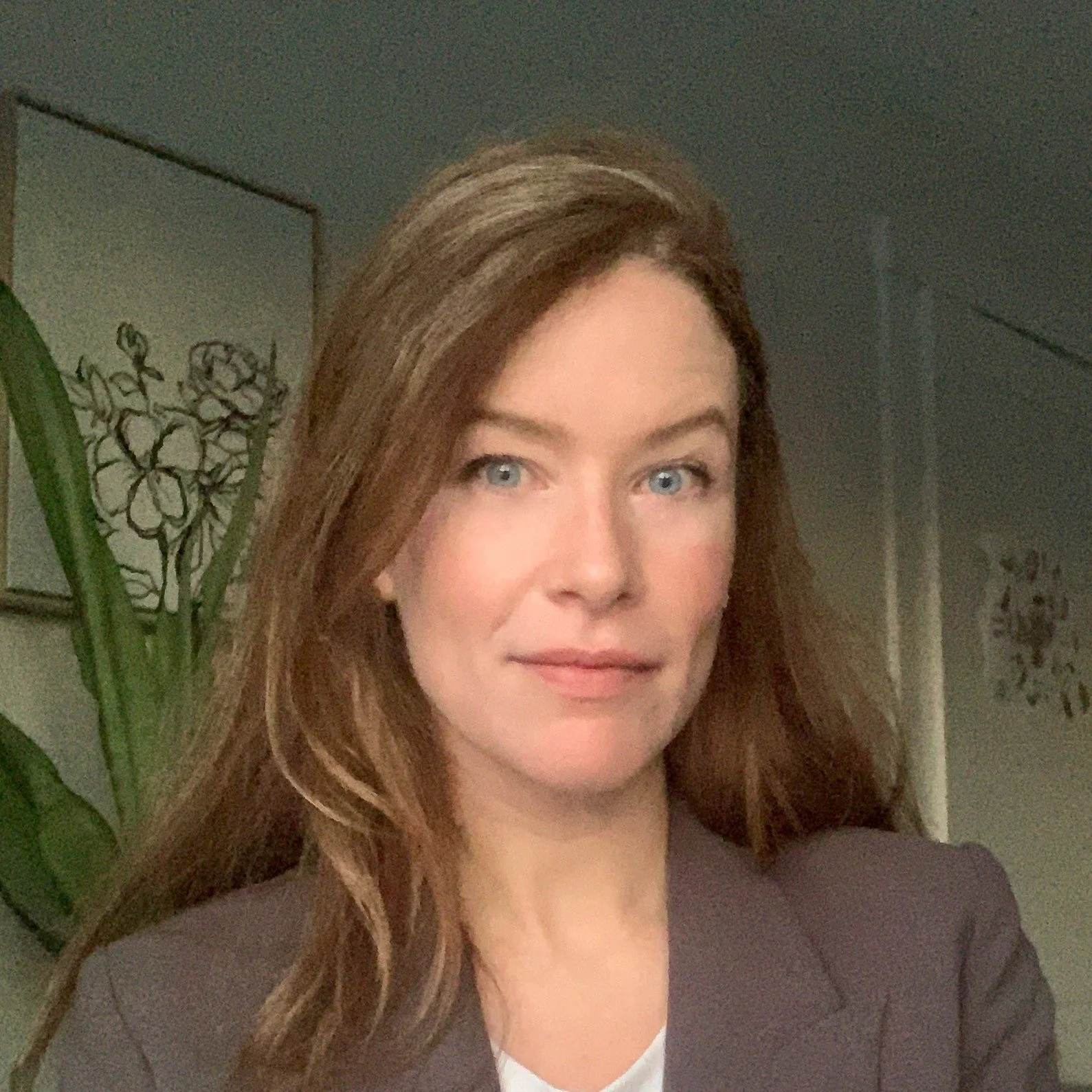 A woman with long brown hair, wearing a gray blazer, stands in front of a wall with framed artwork. A plant is visible in the background.