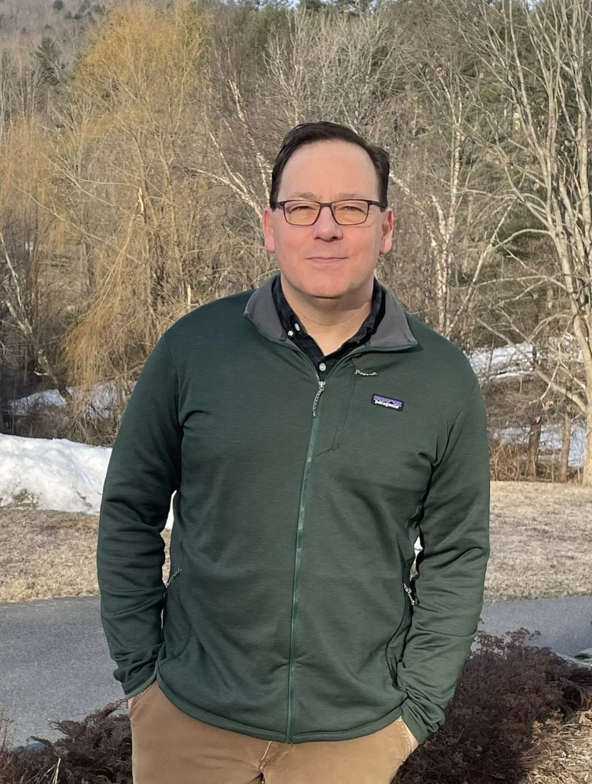 A man wearing glasses and a dark green jacket stands outdoors, with a snowy and wooded background.