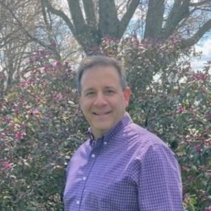 Man smiling in front of a flowering shrub and trees